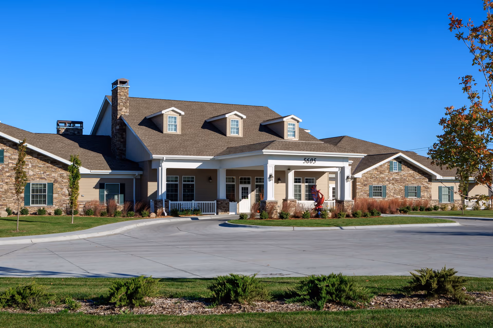 Exterior view of a single-story senior living facility building with a stone and beige siding facade, multiple windows, a covered entrance with white columns, and a driveway in front. The sky is clear and blue, and there is landscaping with grass, bushes, and small trees around the building.