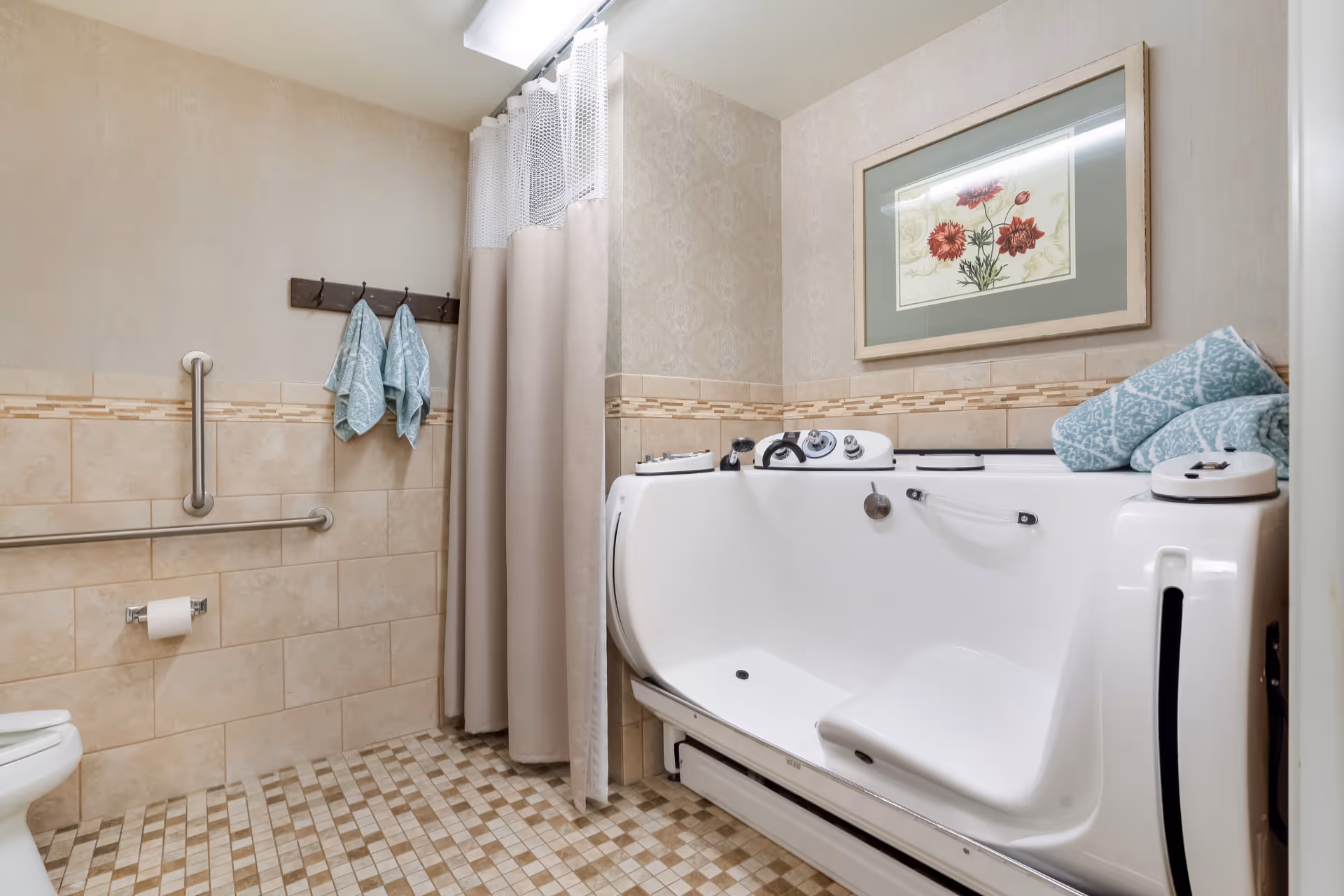 A bathroom with beige tiled walls and floor, featuring a walk-in bathtub with control knobs and a seat. There are two blue patterned towels hanging on a wooden rack, a toilet paper holder with a roll, and a framed floral artwork on the wall above the bathtub. A beige shower curtain is partially drawn beside the bathtub.