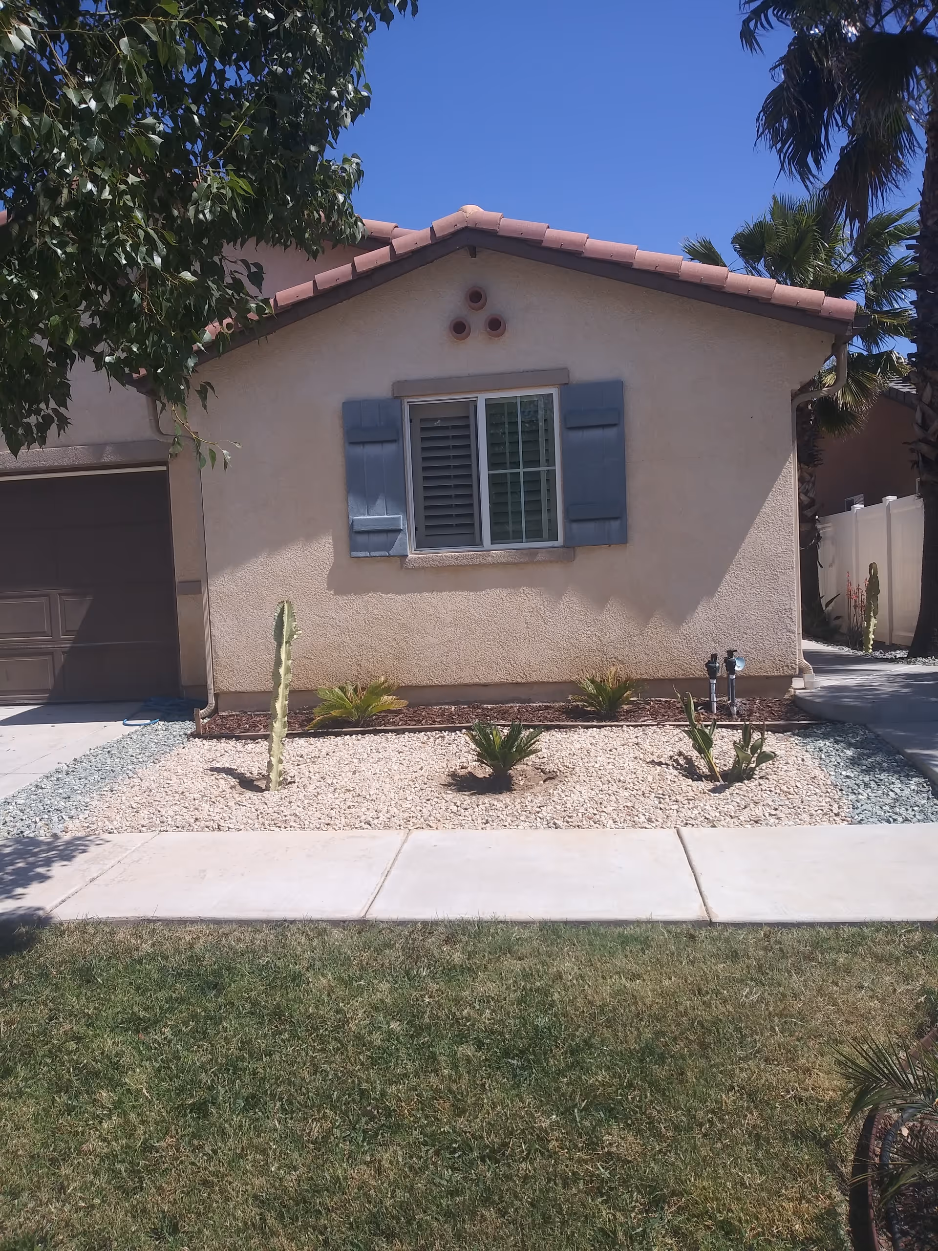 Front exterior of a single-story house with blue shutters, a small rock garden with cacti, a garage to the left and a sidewalk and lawn in front.