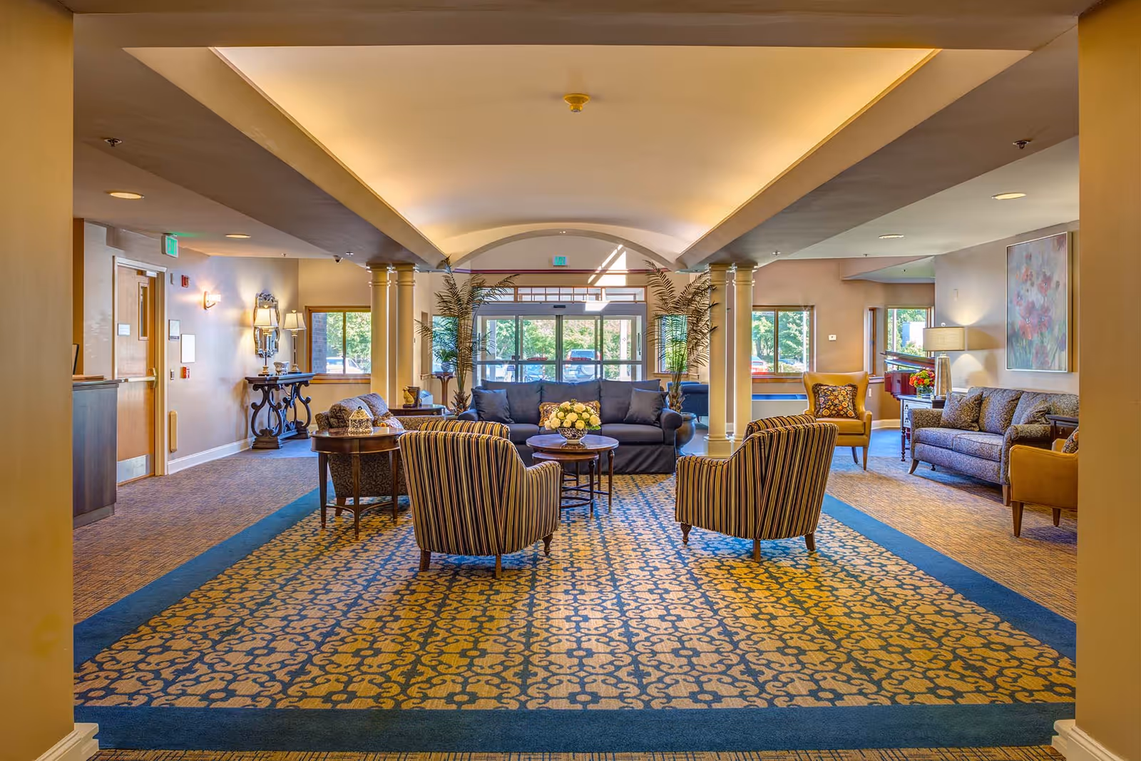 A spacious and well-lit common area with patterned carpet, several seating arrangements including striped armchairs, sofas, and side tables with floral centerpieces. Large windows and glass doors at the far end allow natural light to fill the room. Decorative plants and artwork adorn the space, creating a welcoming atmosphere.