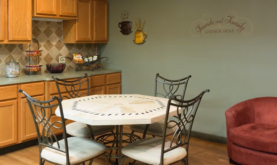 A dining area with a hexagonal table surrounded by four metal chairs with cushioned seats. Behind the table is a countertop with wooden cabinets above and below, holding various decorative items and snacks. On the wall, there is a decorative sign that reads 'Friends and Family GATHER HERE.' A red upholstered chair is partially visible on the right side.