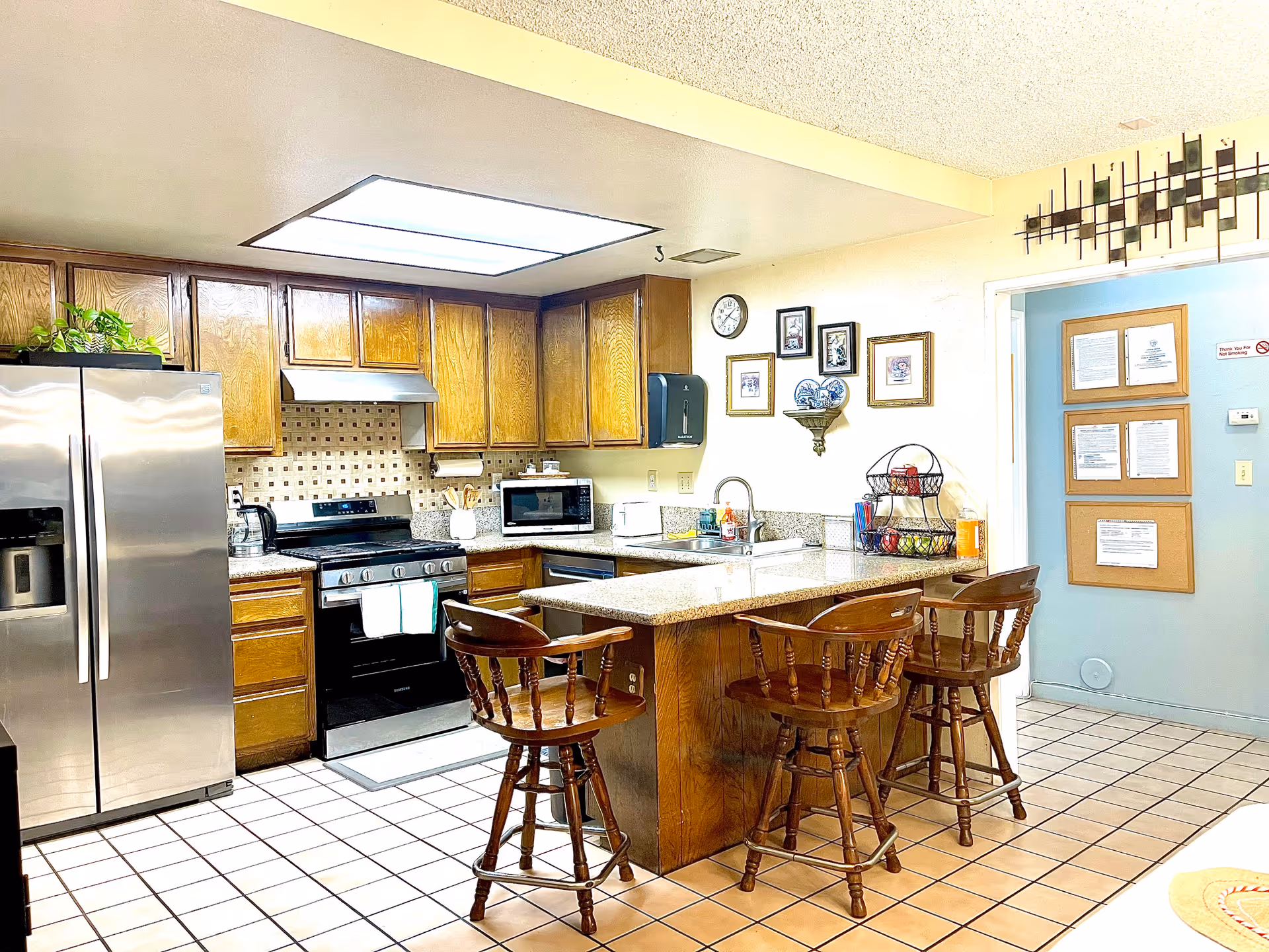 A well-lit kitchen with wooden cabinets, a stainless steel refrigerator, a black stove with a range hood, a microwave, and a granite countertop island with three wooden bar stools. The walls have framed pictures and a clock, and there is a basket with fruit on the counter. The floor is tiled with a mix of white and brown tiles.
