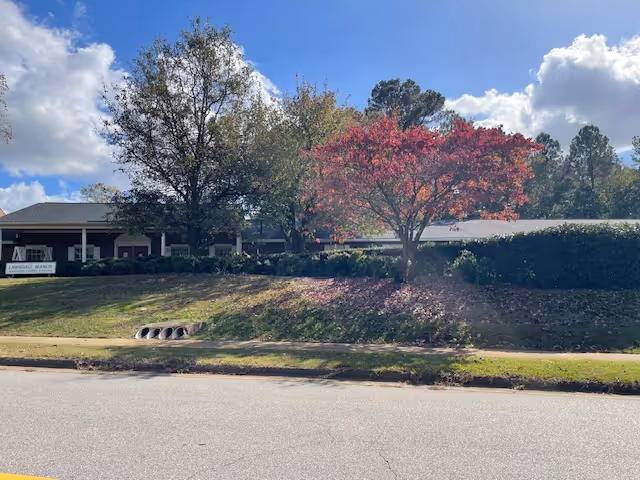 Exterior view of Lawndale Manor Assisted Living facility with a grassy front yard, several trees including one with red leaves, a sidewalk, and a clear blue sky with some clouds.
