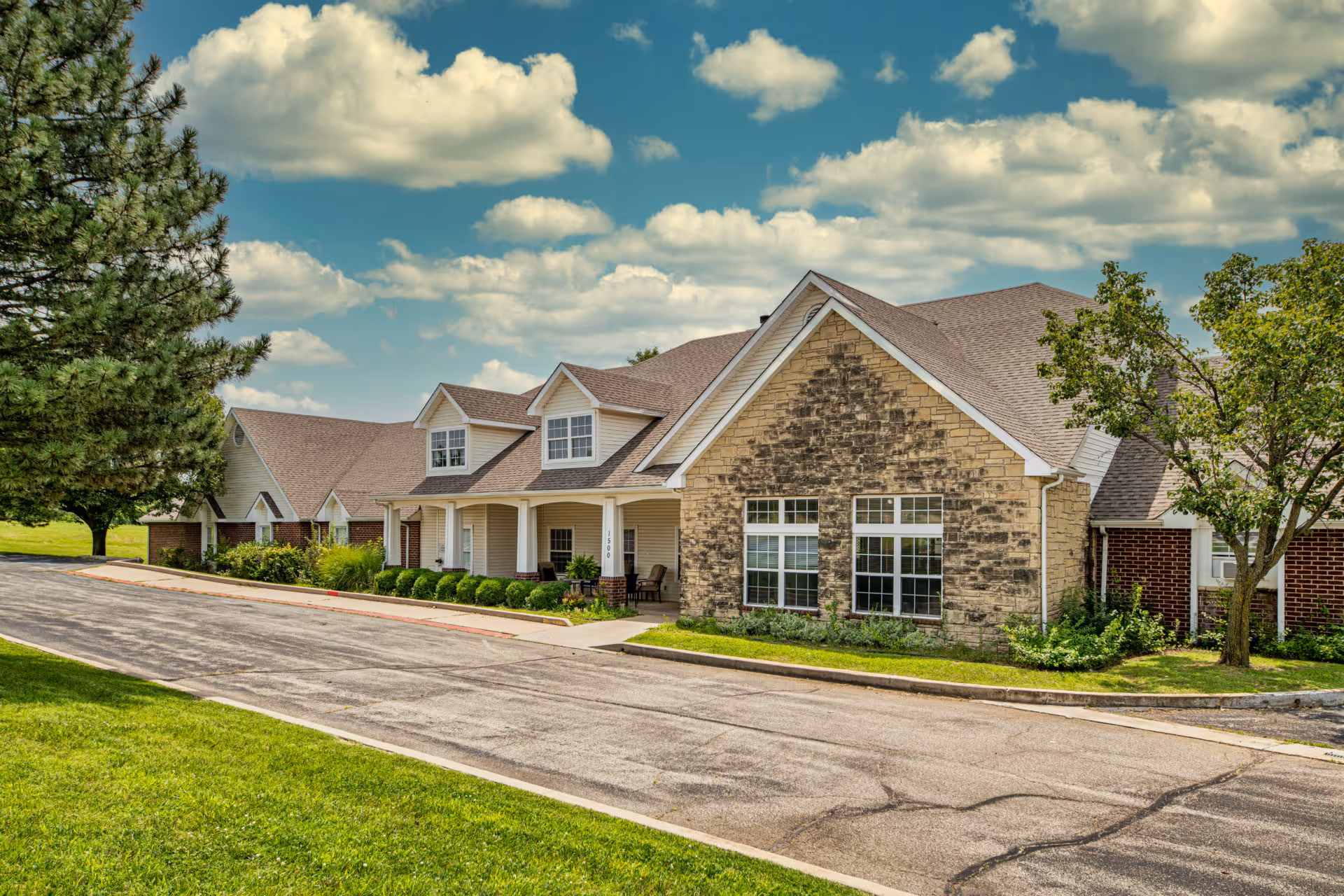 Exterior view of Creekside Village facility showing a single-story building with a stone and brick facade, multiple windows, a covered porch with chairs, surrounded by green grass, trees, and a partly cloudy sky.