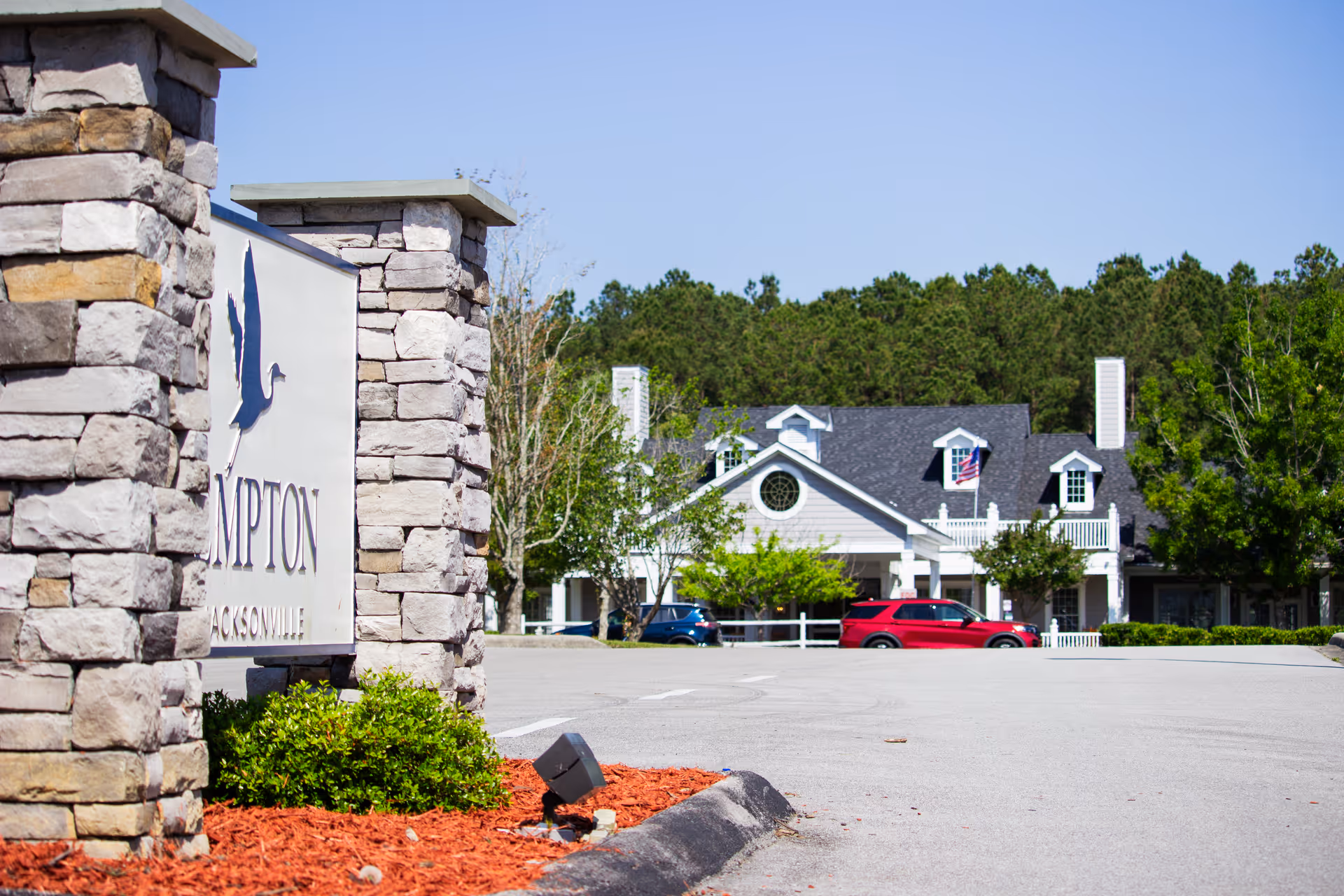 Stone entrance sign and landscaped driveway leading to a two-story white building with parked cars and trees.