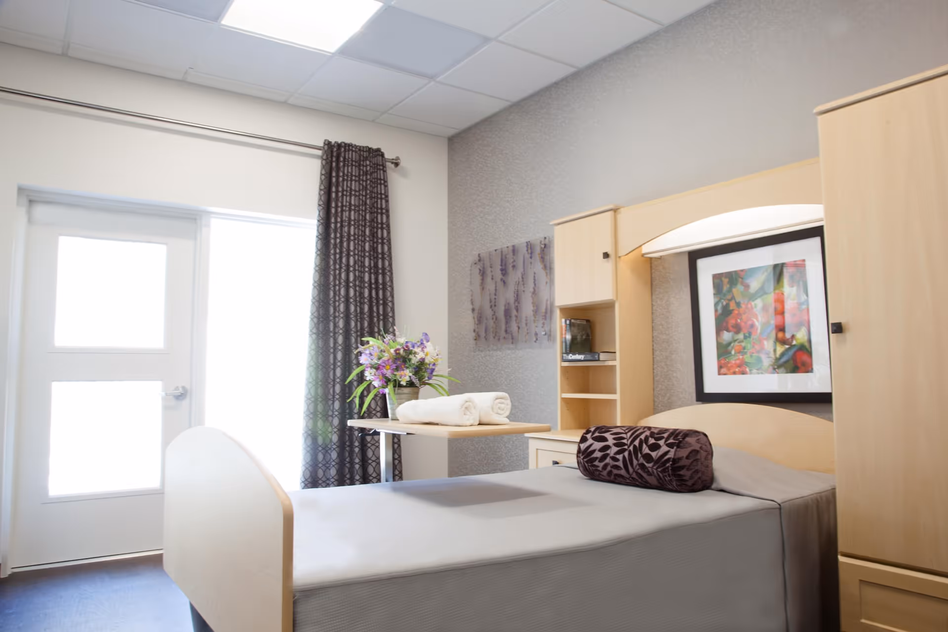 A bright and clean bedroom in a senior living facility with a single bed covered in a gray sheet and a decorative purple bolster pillow. The room features light wood furniture including a headboard with built-in shelves and cabinets. A small table next to the bed holds a vase with purple flowers and two rolled white towels. There is a large window or door with sheer white curtains and dark patterned drapes, and a colorful framed artwork hangs on the wall above the bed.