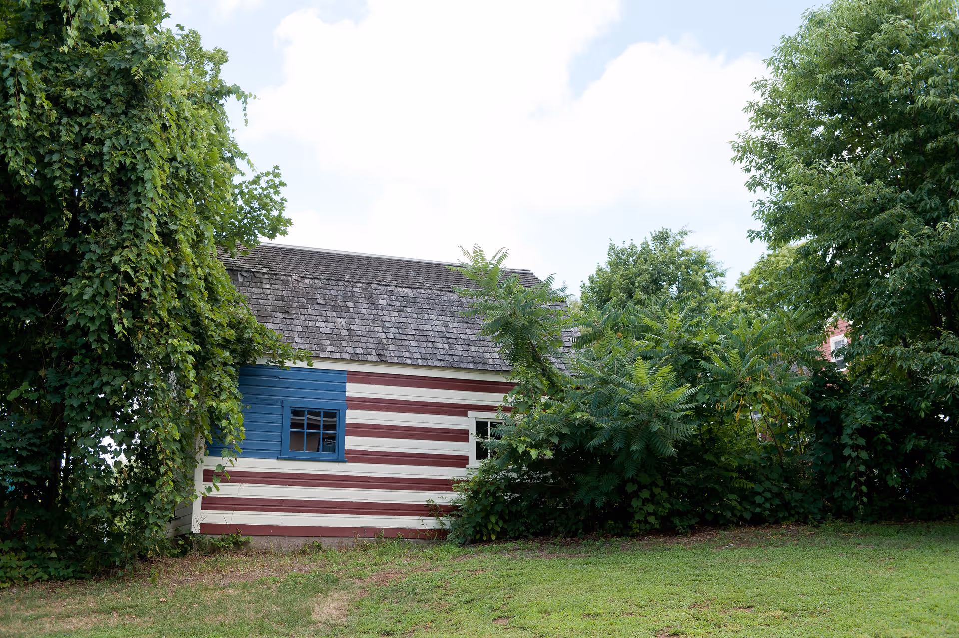 Small wooden building painted with red and white horizontal stripes and a blue window area, partially obscured by trees and bushes.