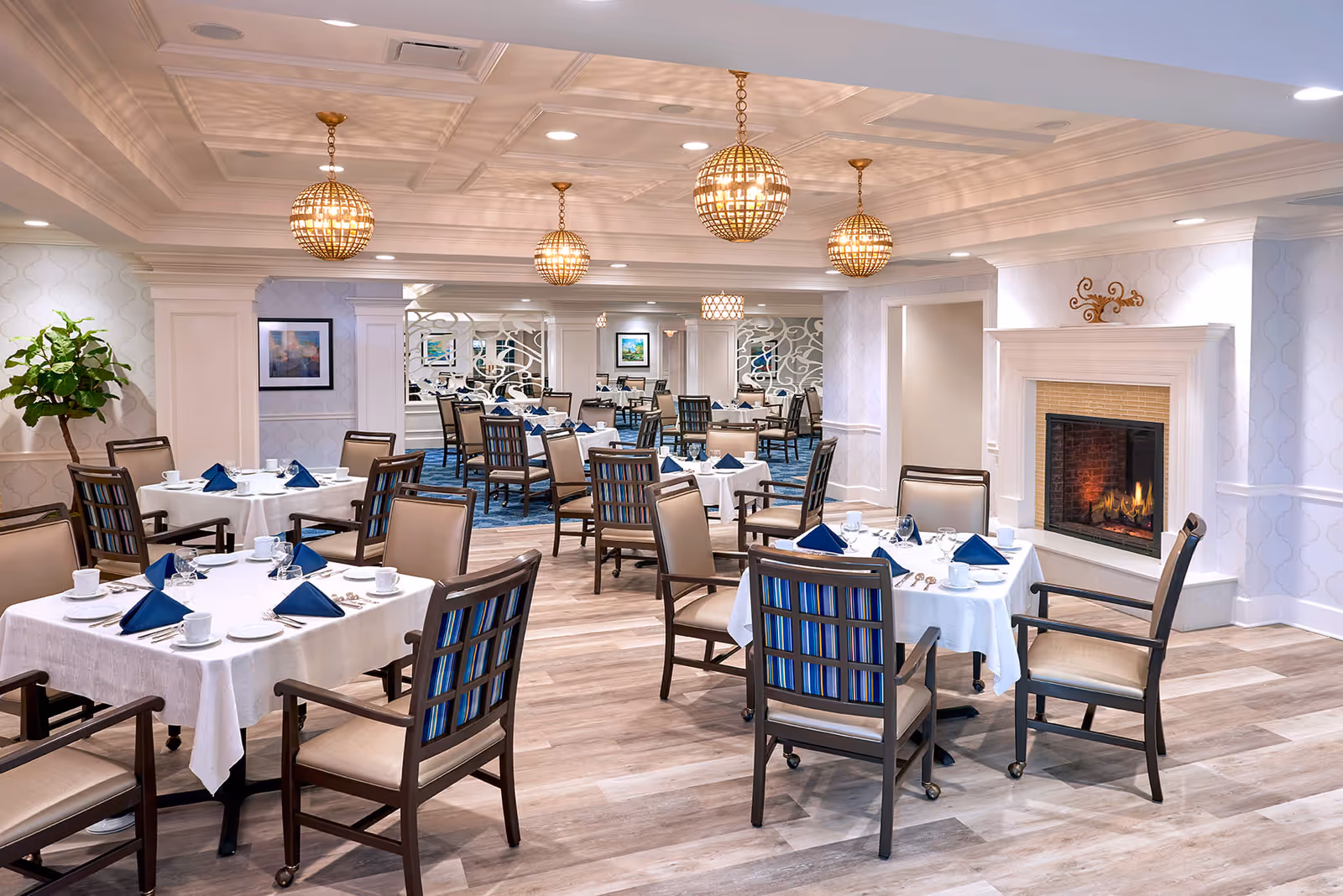Bright, elegant dining room with multiple tables set with white linens and blue napkins, wooden chairs, and a lit fireplace.