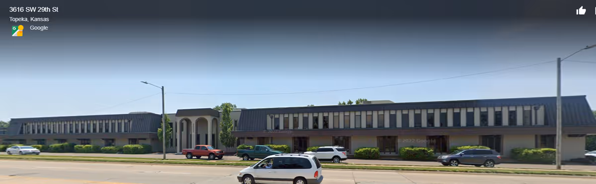 Long, single-story building with a central arched entrance, cars parked along the curb.