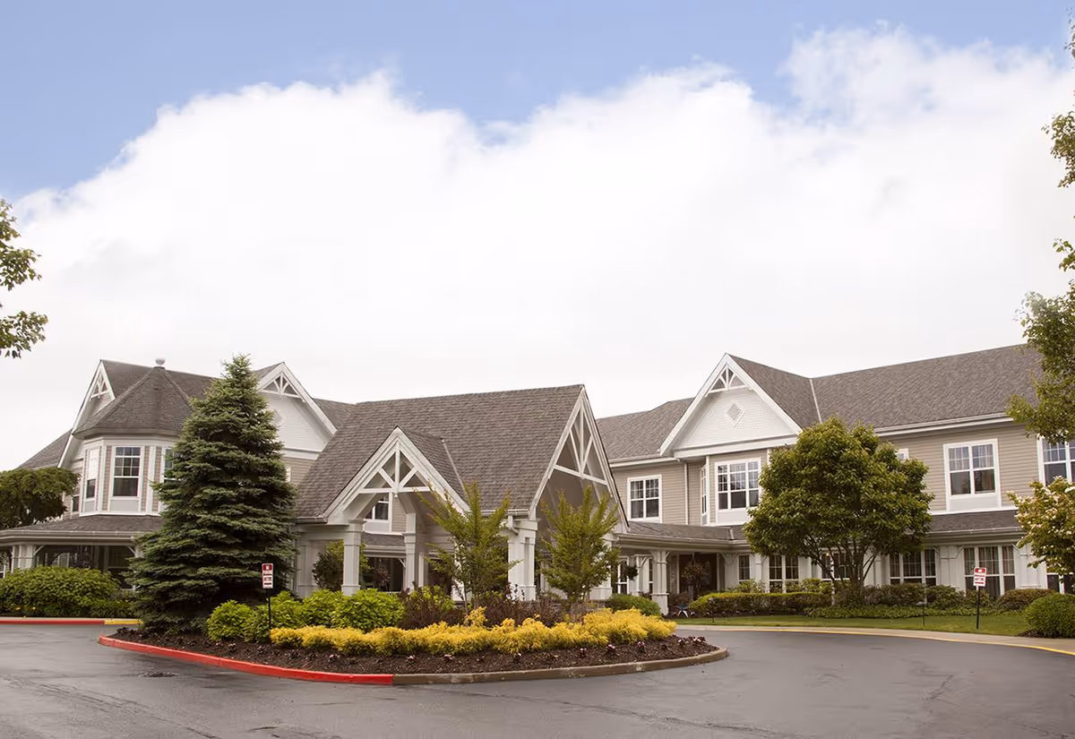 Exterior view of a senior living facility named Sunrise of Holbrook featuring a large building with multiple windows, a peaked roof, and a covered entrance. The driveway is circular with landscaped greenery including bushes, trees, and a large evergreen tree. The sky is partly cloudy.