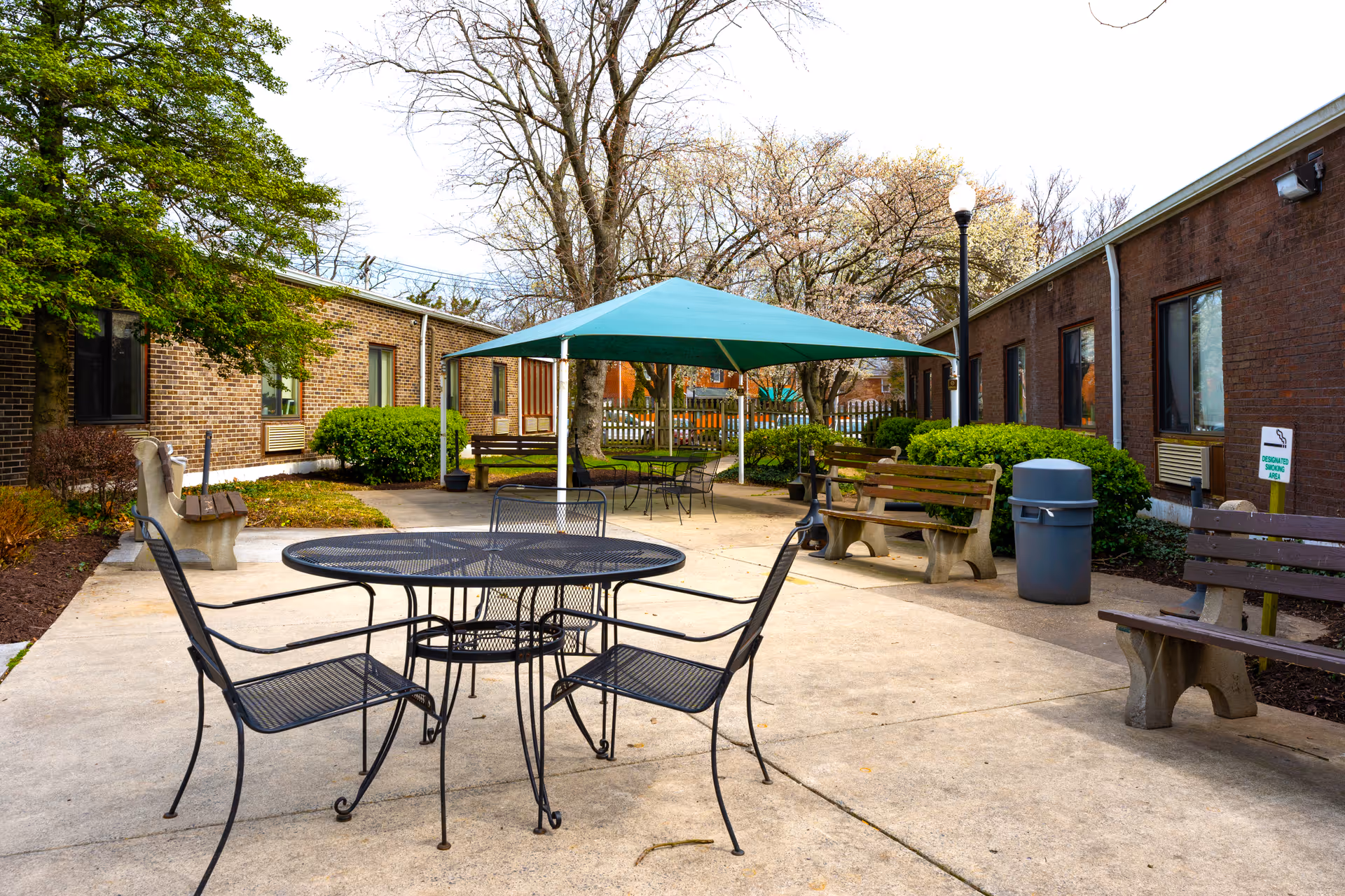 Outdoor patio area at Autumn Lake Healthcare at Homewood Center featuring metal tables and chairs, wooden benches, a large green canopy providing shade, surrounded by brick buildings and greenery.