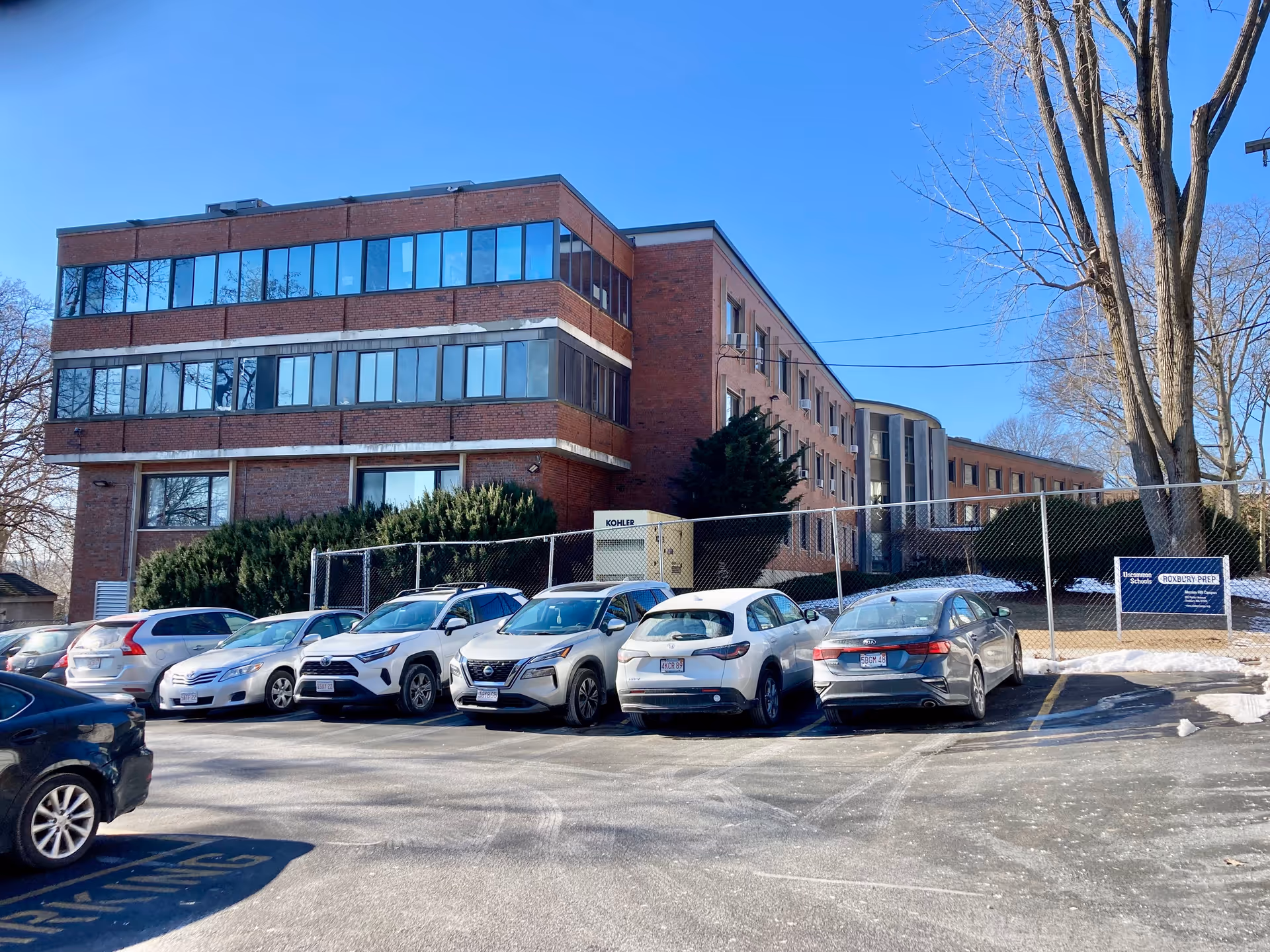 Exterior view of a multi-story brick building with large windows, a parking lot with several parked cars in front, and leafless trees under a clear blue sky.