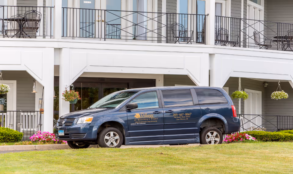 A dark blue Village at Mariner's Point van parked in front of a two-story senior living building with balconies and hanging flower baskets.