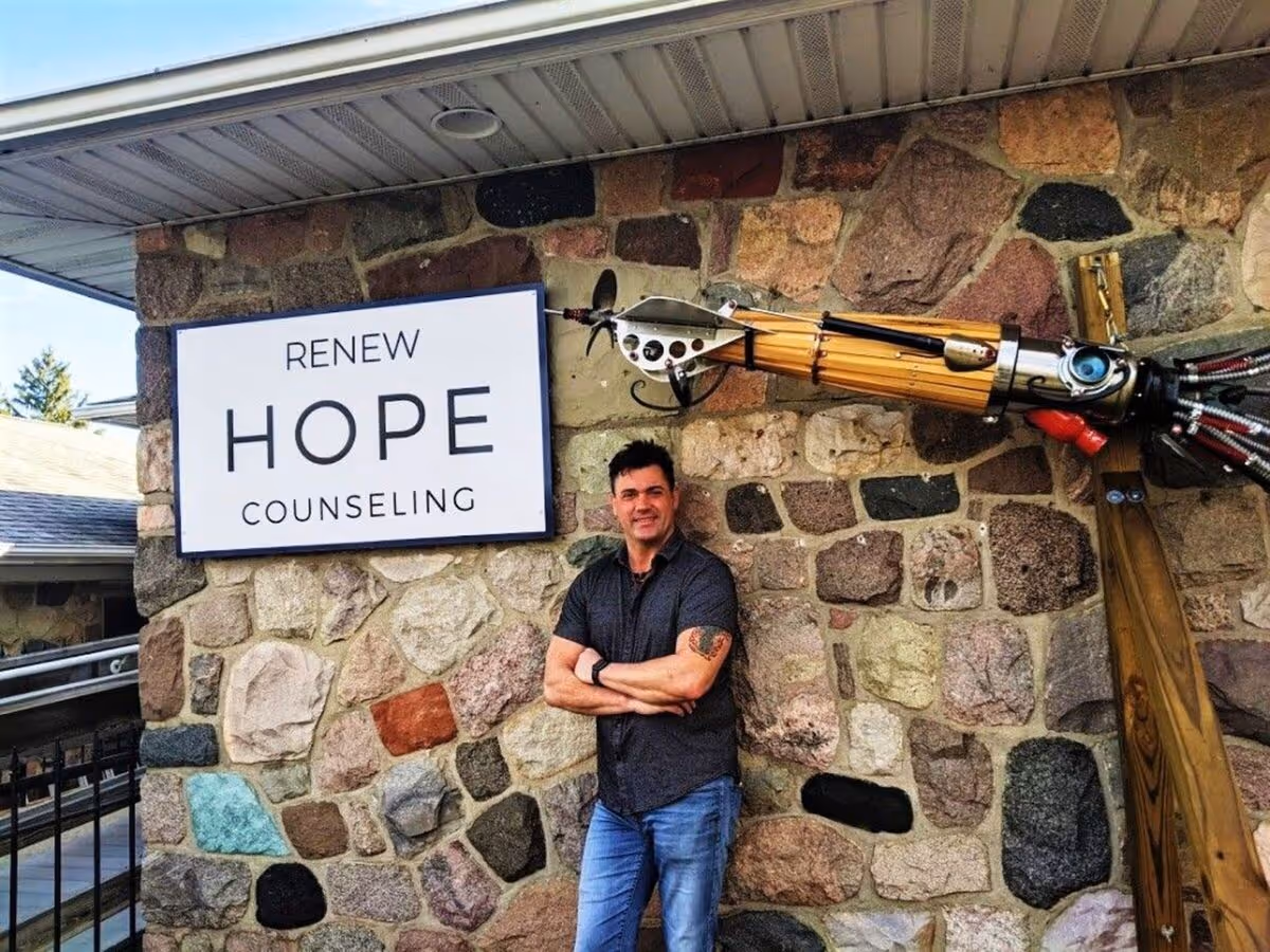 A man stands with arms crossed beside a stone wall and 'Renew Hope Counseling' sign.