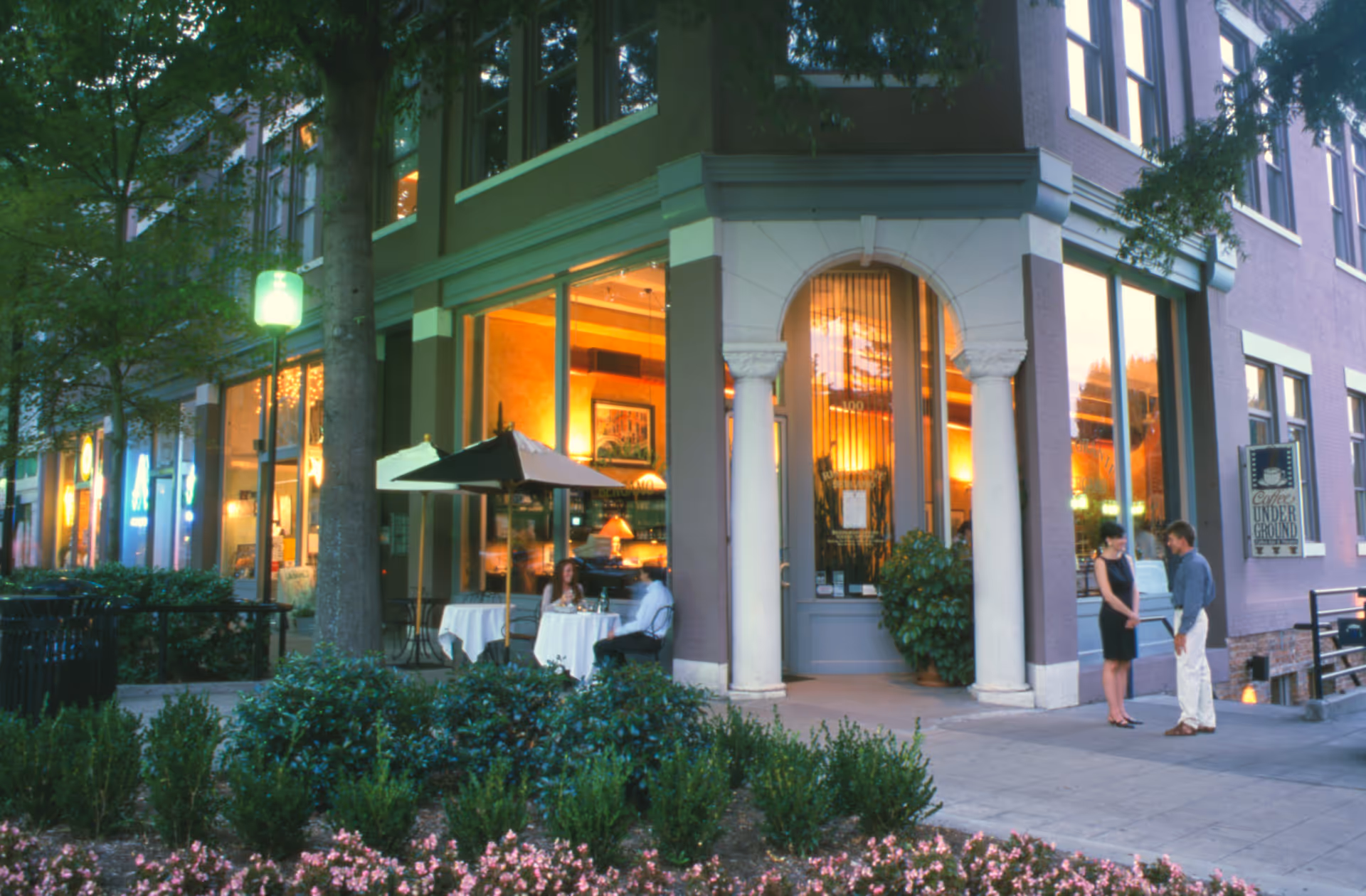 Corner building with lit storefronts, outdoor seating, and two people conversing on the sidewalk.
