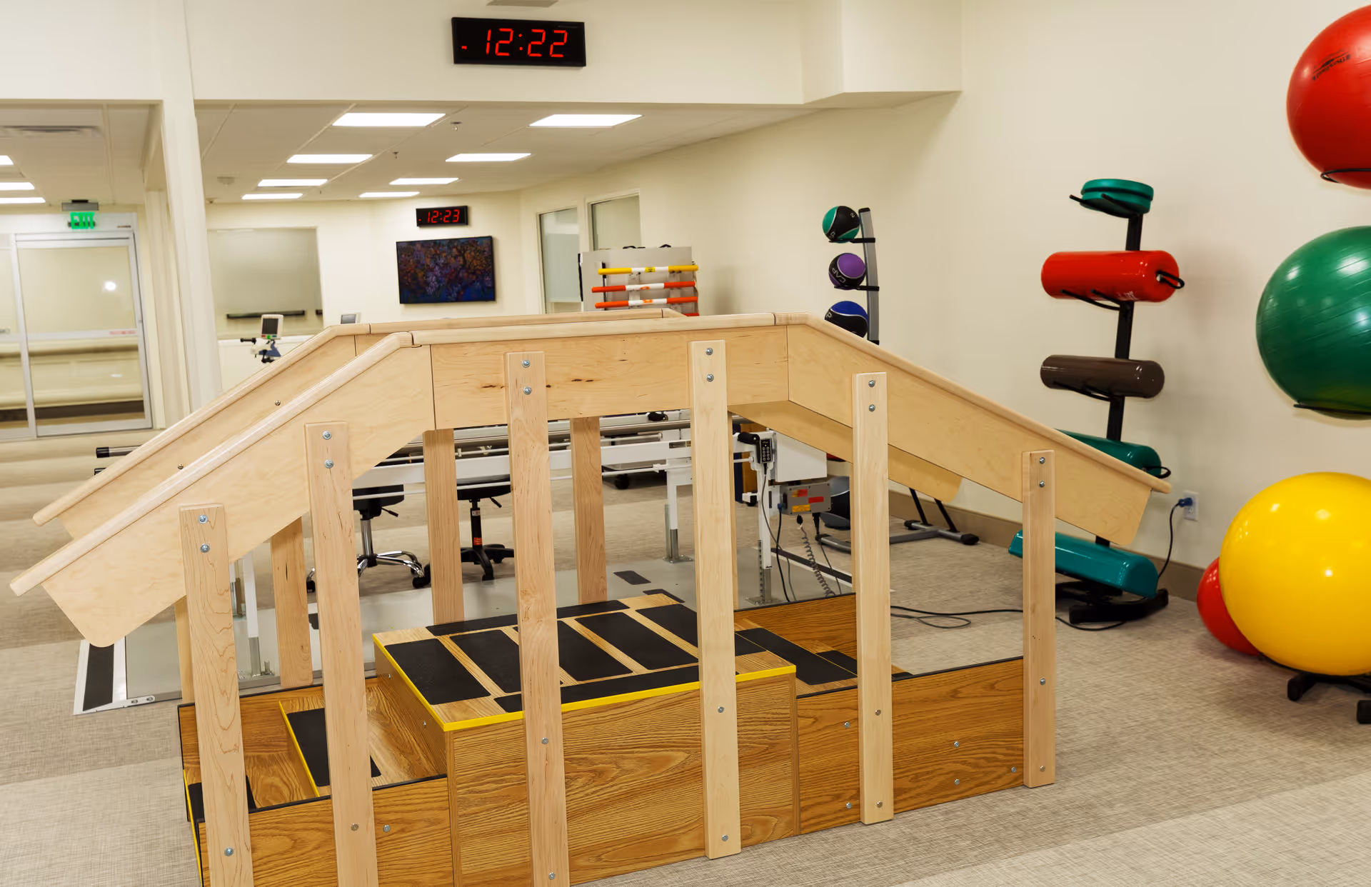 Indoor physical therapy or rehabilitation room with wooden stairs and handrails for mobility exercises. Various exercise balls and equipment are visible on the right side, and a digital clock on the wall shows the time as 12:22.