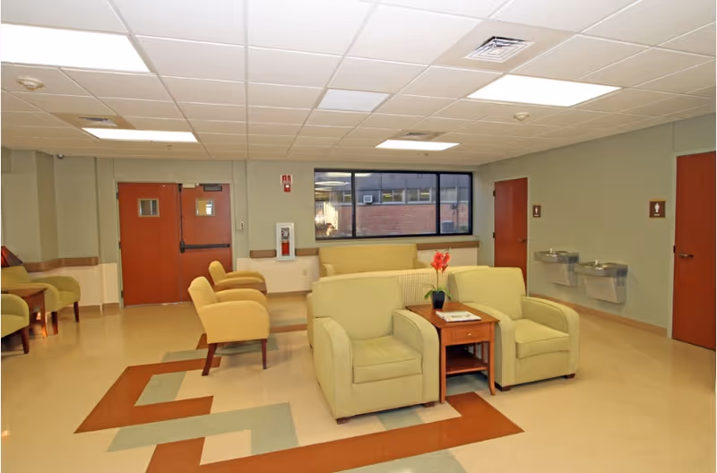 A waiting area with several green armchairs and a small wooden table with a potted plant and magazines. There are two water fountains mounted on the wall to the right, a large window showing a brick building outside, and double doors on the left side of the room. The floor has a geometric pattern in brown and green colors.