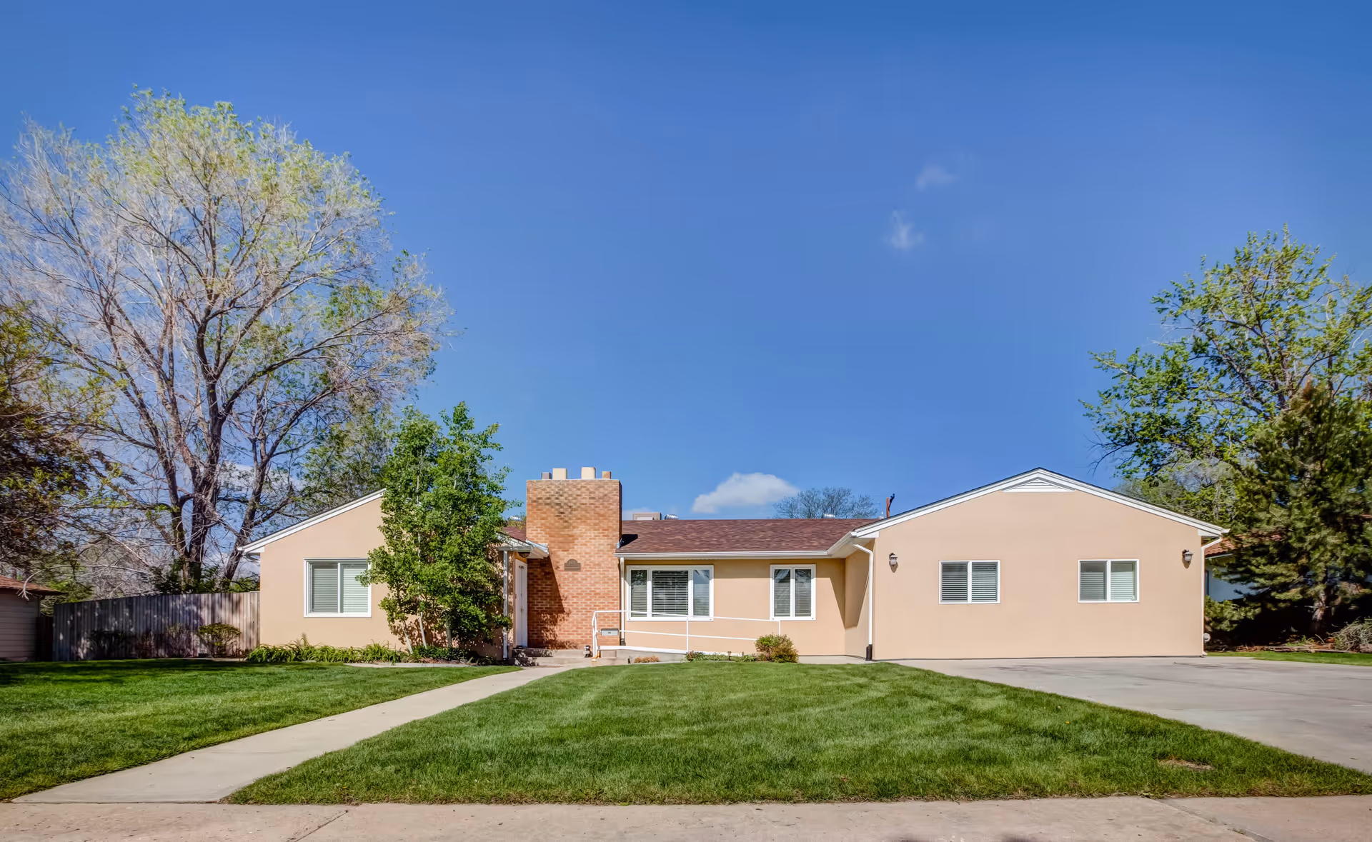 Single-story ranch-style building with tan stucco, a central brick chimney, front lawn, driveway and trees under a clear blue sky.