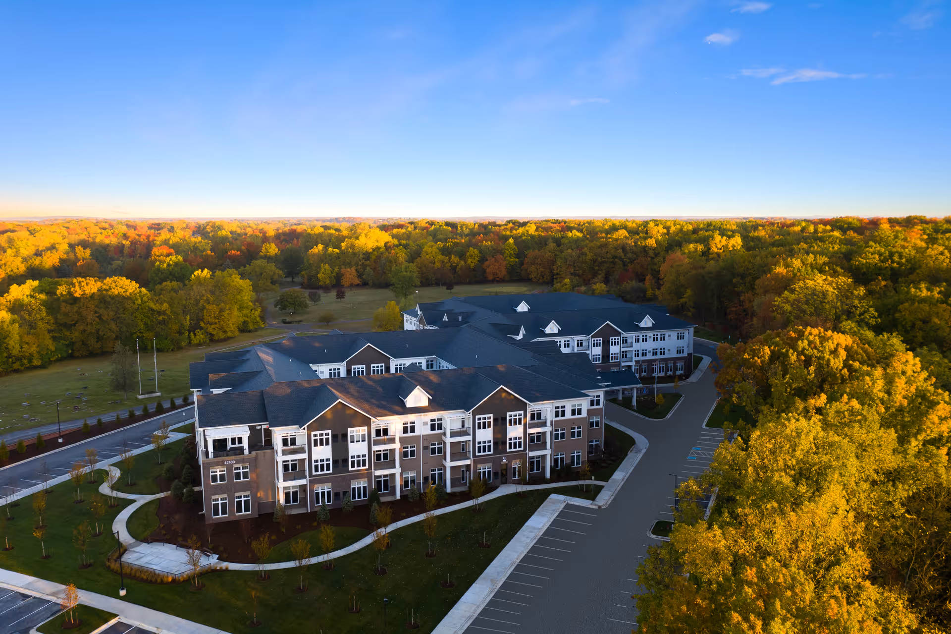 Aerial view of a large senior living facility building surrounded by trees with autumn foliage under a clear blue sky. The building has multiple stories with numerous windows and a parking area in front.
