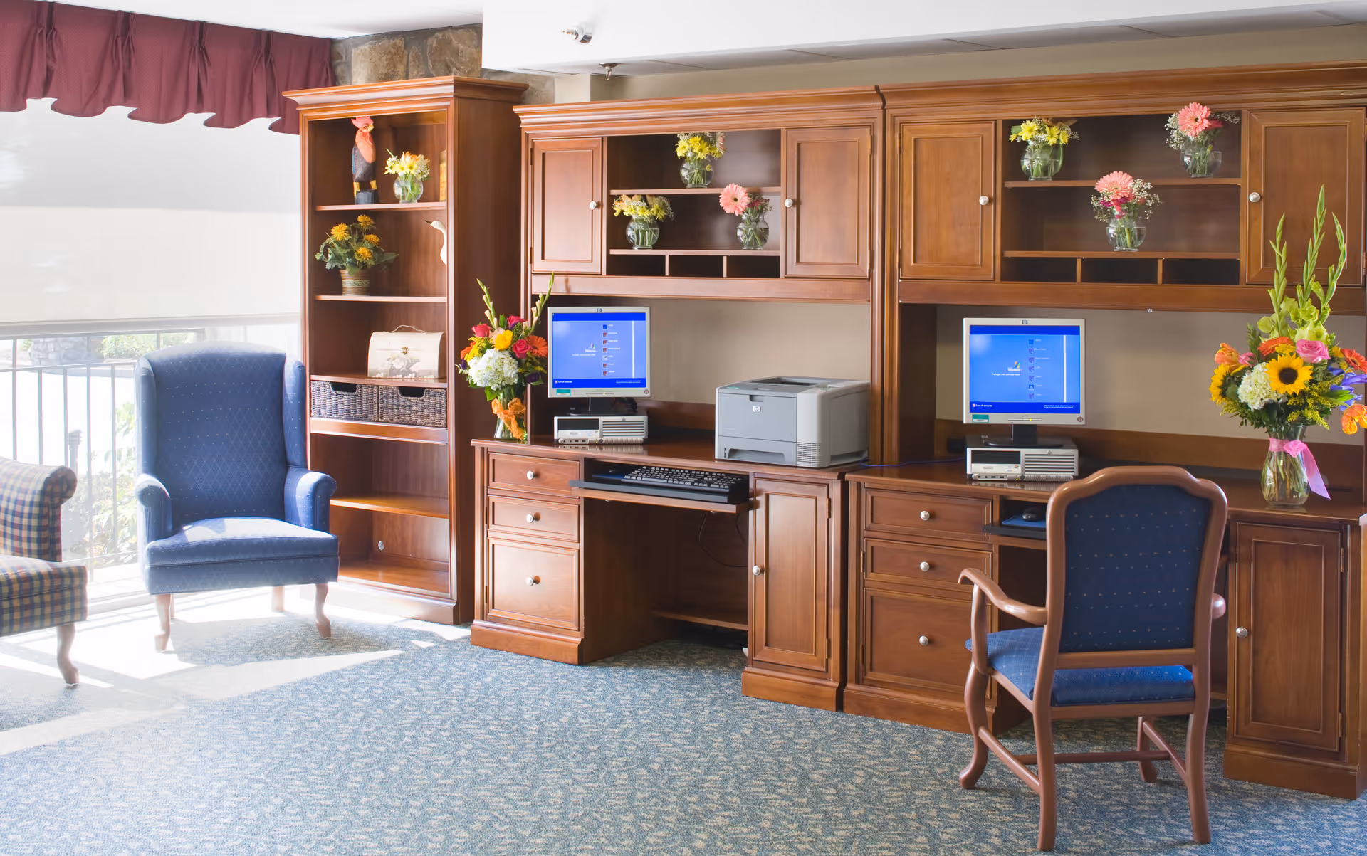A bright room with two wooden desks featuring desktop computers and a printer. The desks have upper cabinets and shelves decorated with vases of colorful flowers. There are two blue upholstered chairs, one with armrests and one wingback style, positioned near the desks. A large window with a maroon valance lets in natural light, illuminating the carpeted floor.