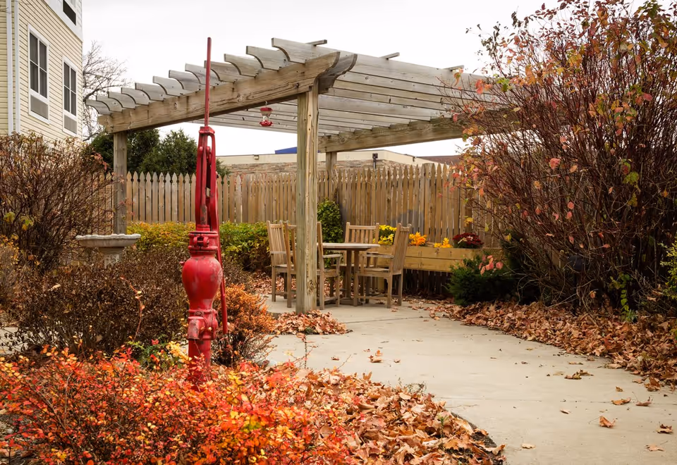 Courtyard with a wooden pergola, outdoor table and chairs, a red hand pump, and fallen autumn leaves.