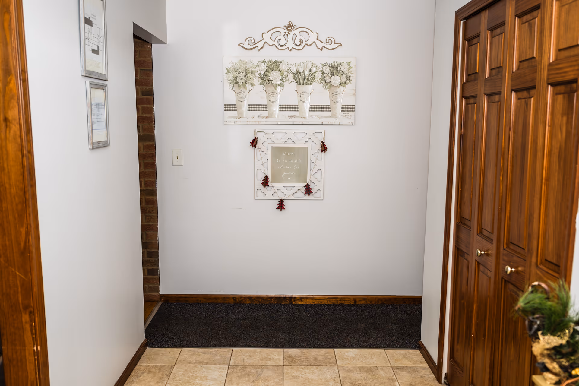 A hallway with white walls and tiled floor leading to a dark carpeted area. On the right side, there are wooden closet doors with brass knobs. On the left wall, two framed pictures are hung. On the far wall, there is a decorative wall hanging with floral artwork and a smaller framed quote with small red decorations hanging from it.