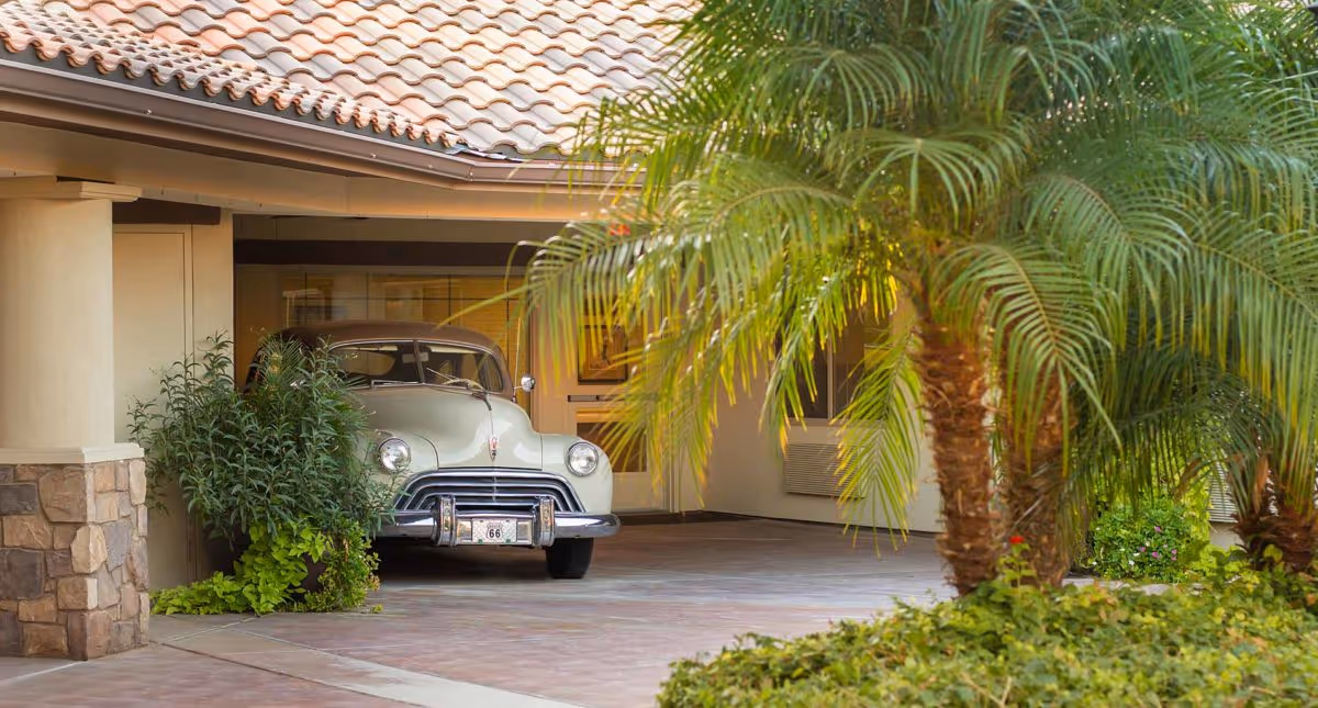 A vintage car parked under a covered entrance surrounded by palm trees and landscaping at a building front.