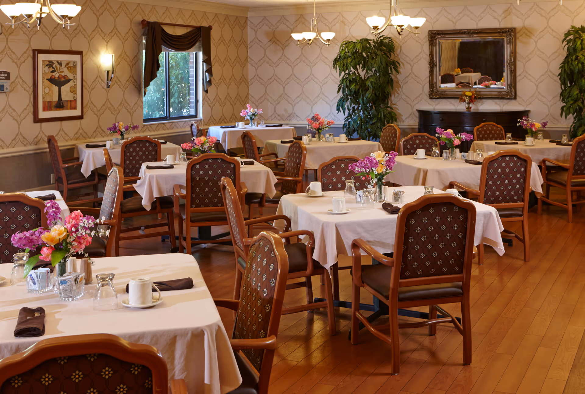 A dining room with multiple tables covered in white tablecloths, each set with cups, glasses, and napkins. The room features wooden chairs with patterned upholstery, floral centerpieces on each table, hardwood flooring, patterned wallpaper, a window with a dark curtain, a large mirror on the wall, and two large potted plants.