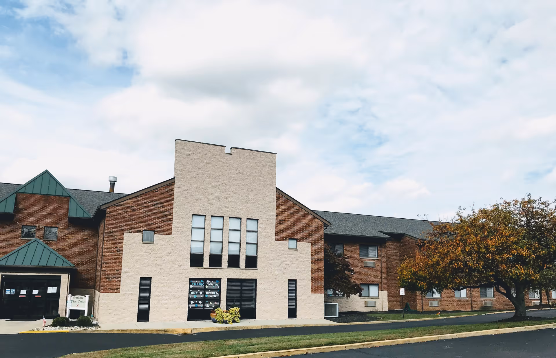 Exterior view of a two-story brick building with a beige central section and green roof accents under a partly cloudy sky. There is a tree with autumn-colored leaves on the right side and a paved driveway in front. A sign near the entrance reads 'The Oaks'.