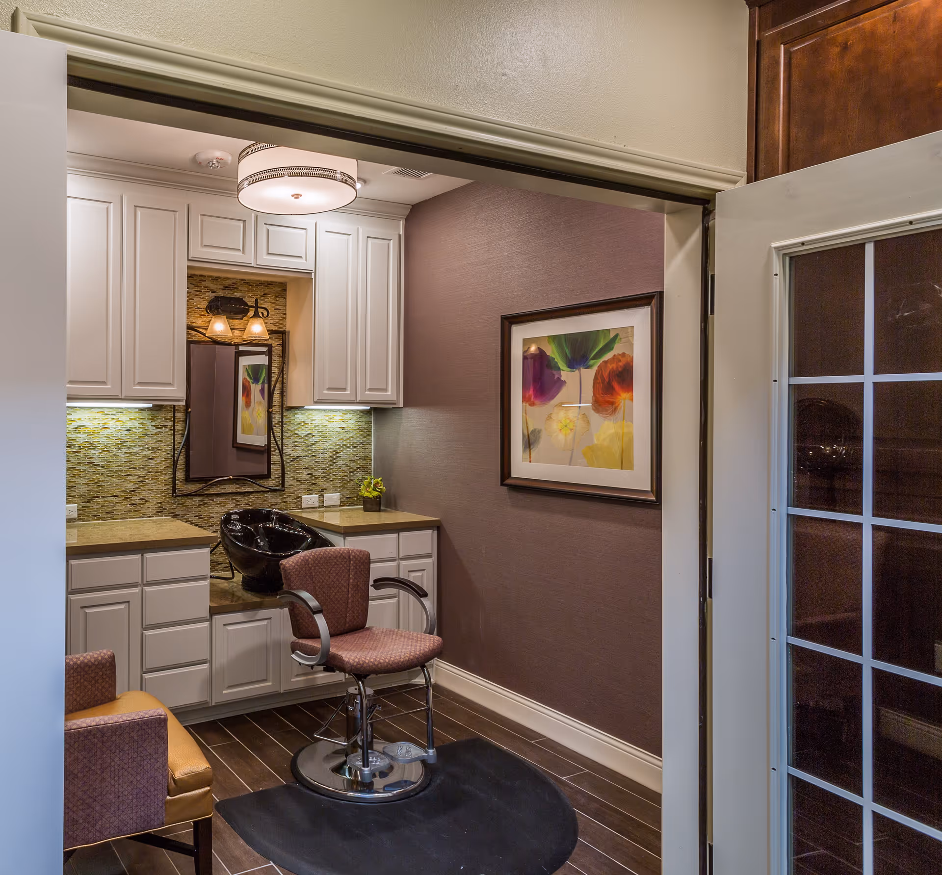 Interior view of a small hair salon area within a facility, featuring a salon chair in front of a black wash basin, white cabinetry with under-cabinet lighting, a mirror with two wall-mounted lights above it, a framed colorful floral artwork on a purple wall, and a glass-paneled door partially open to the right.