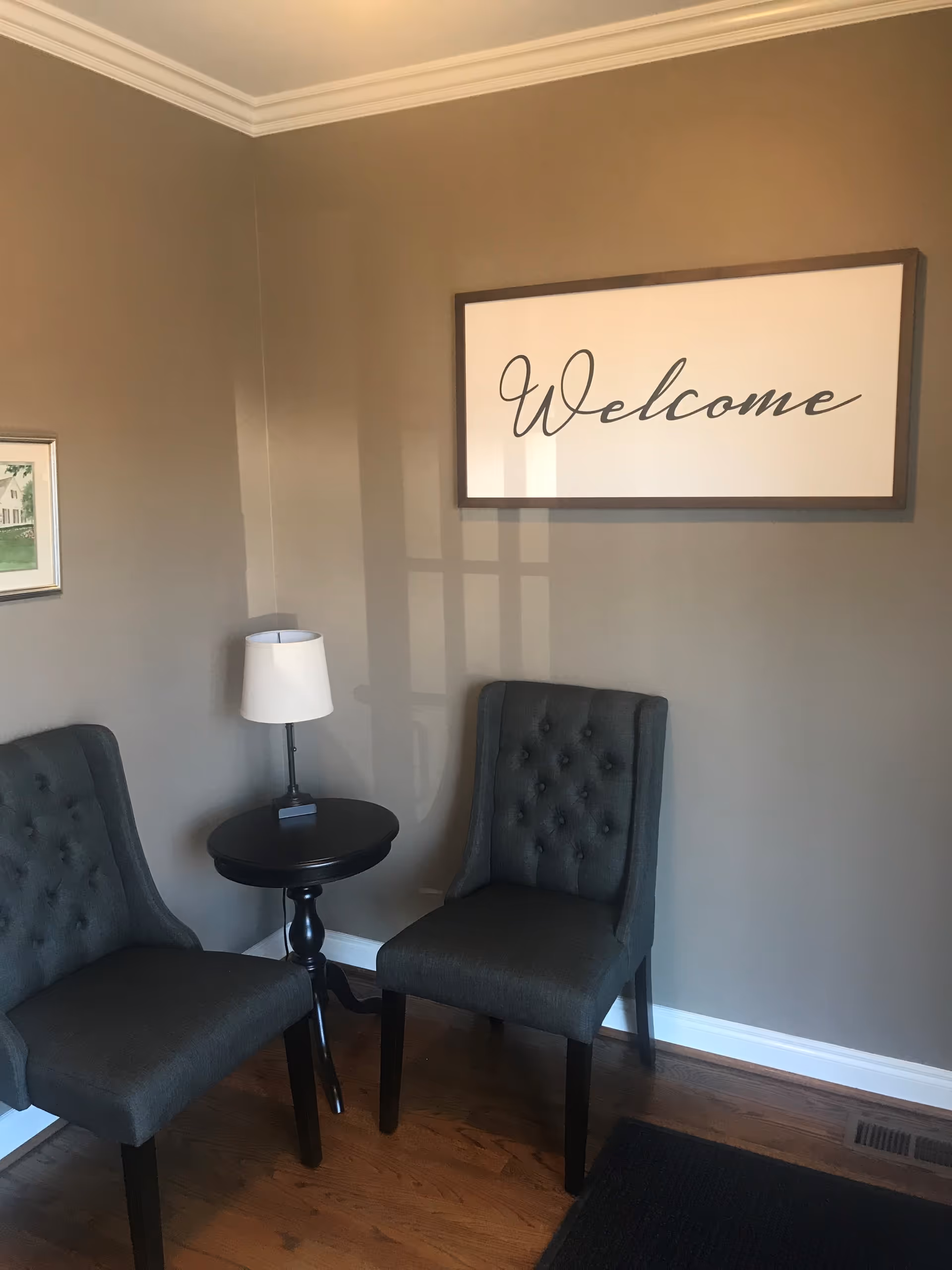 Corner waiting area with two gray upholstered chairs, a round table, lamp, and Welcome sign.
