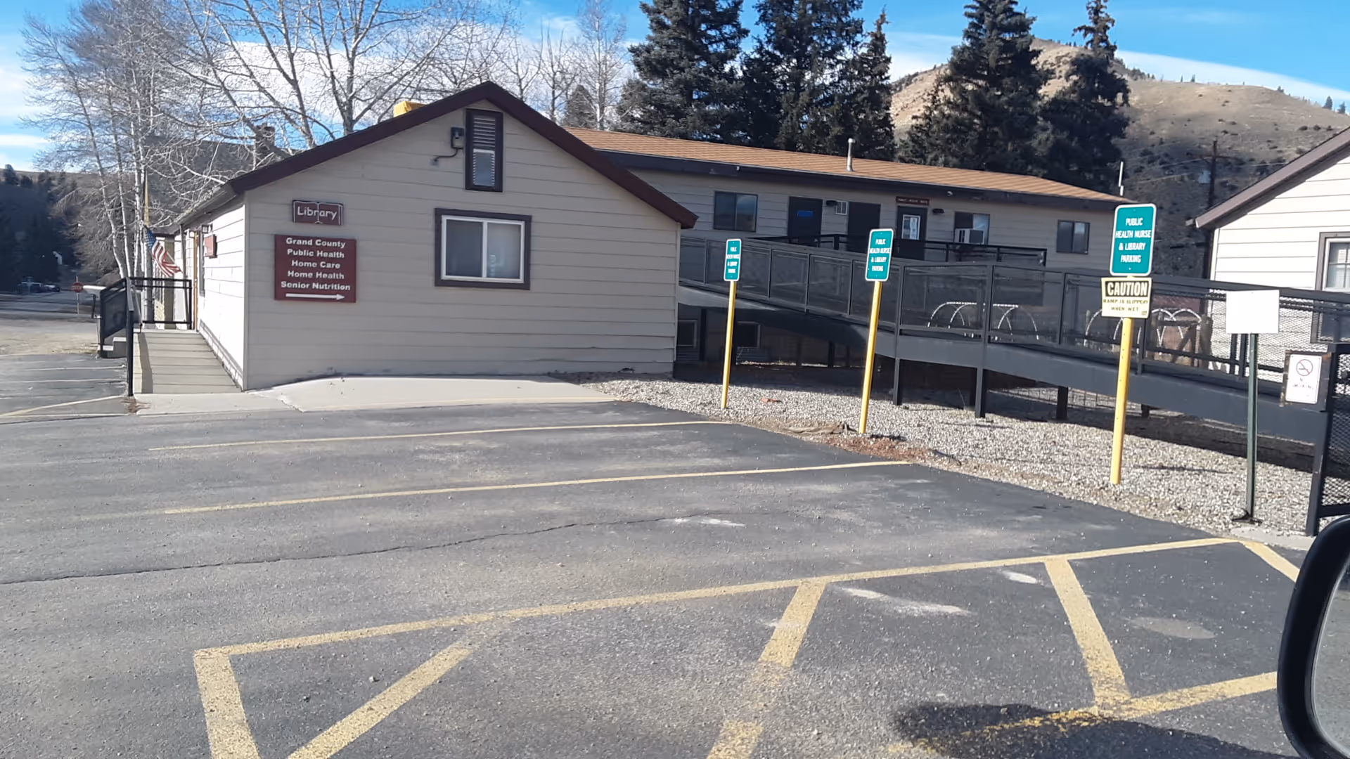 Exterior view of a small beige building with ramp, signage, parking lot, and surrounding trees.