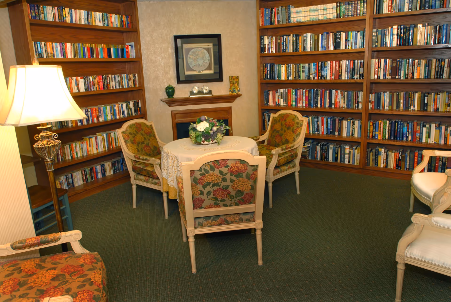 Cozy library room with wooden bookshelves filled with books along two walls, a small round table covered with a lace tablecloth and a floral centerpiece, surrounded by four upholstered chairs with floral patterns. A floor lamp is on the left side, and a framed map hangs above a small wooden mantelpiece.