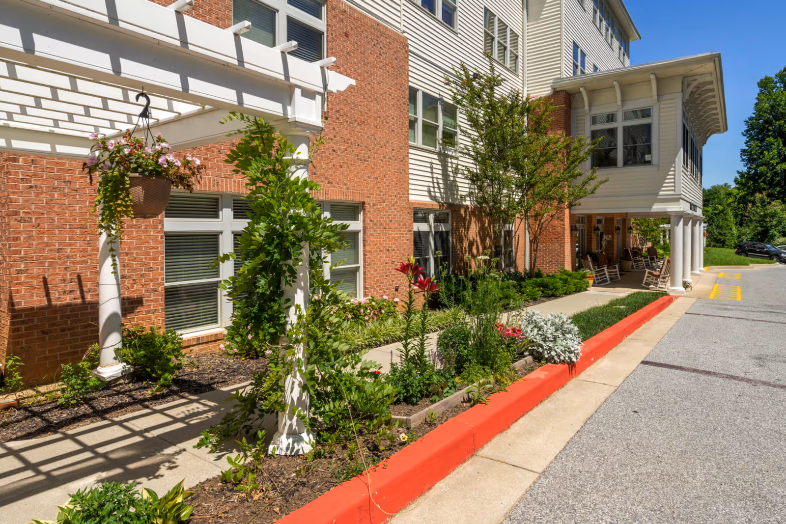 Front entrance of a senior living building with a pergola, landscaped flower beds and a covered porch with rocking chairs.
