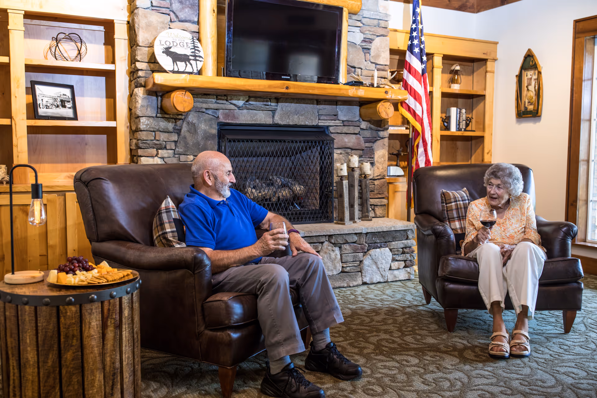 An elderly man and woman sitting in leather armchairs in a cozy living room with a stone fireplace and wooden shelves. The man is wearing a blue shirt and gray pants, holding a drink, while the woman is wearing a floral blouse and white pants, holding a glass of wine. There is an American flag beside the fireplace and a small round wooden table with a plate of cheese and grapes.