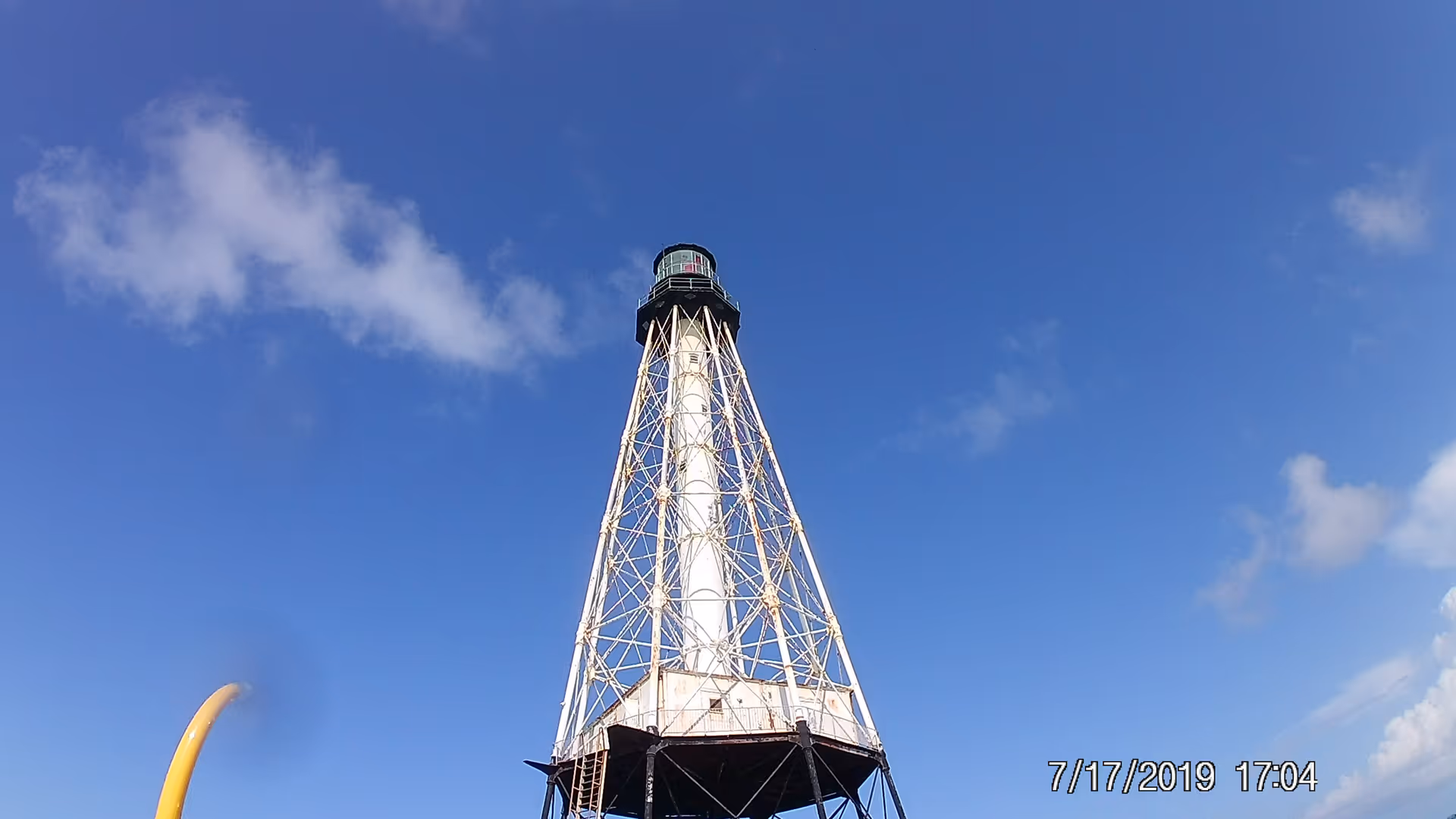 Tall white lattice lighthouse tower against blue sky with a date stamp.
