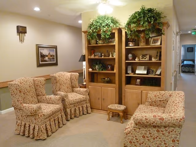 Cozy senior living common area with floral upholstered chairs, a tall bookshelf, and plants.