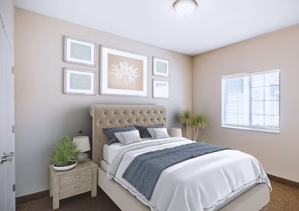 A bright bedroom with a tufted upholstered bed, bedside table, framed artwork, and a window.