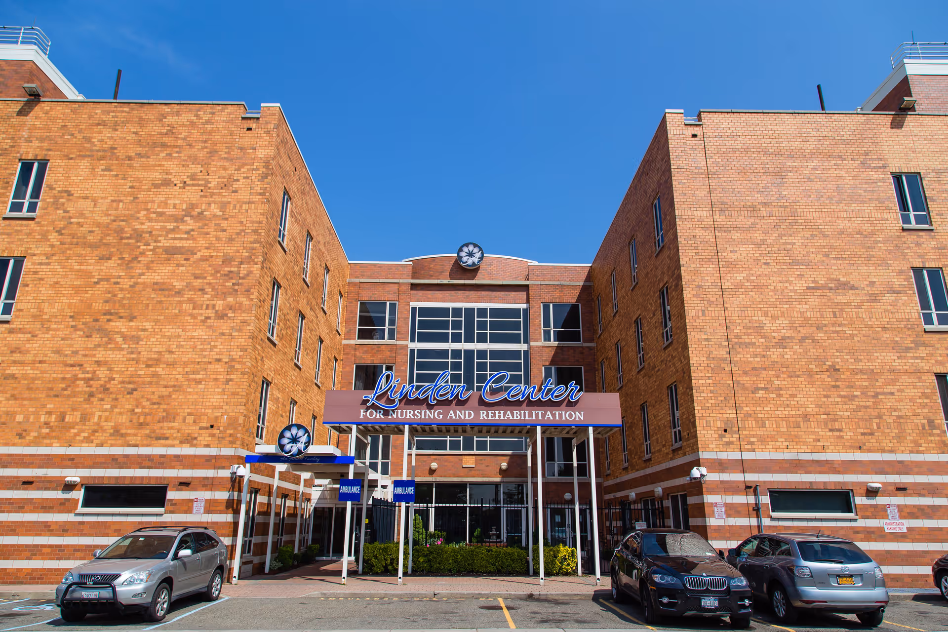 Front exterior view of the Linden Center for Nursing and Rehabilitation, a multi-story brick building with large windows and a covered entrance. Several cars are parked in front under a clear blue sky.