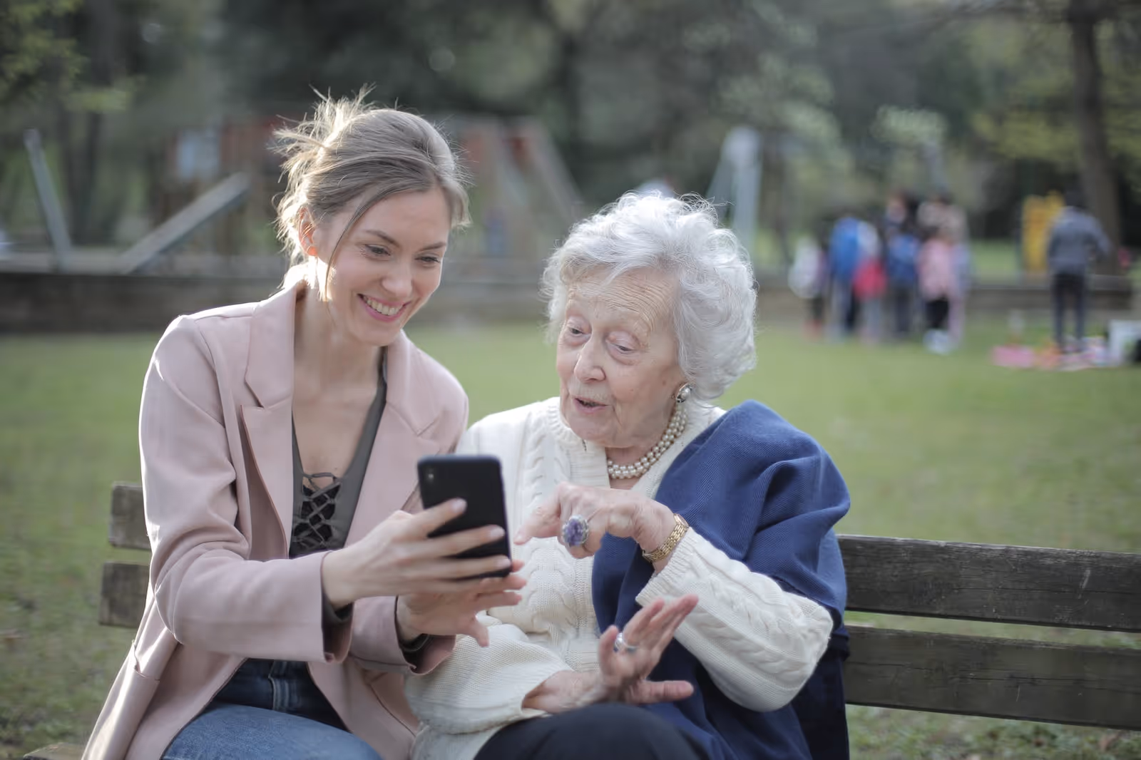 An elderly woman and a younger caregiver sit on a park bench, looking at a smartphone.