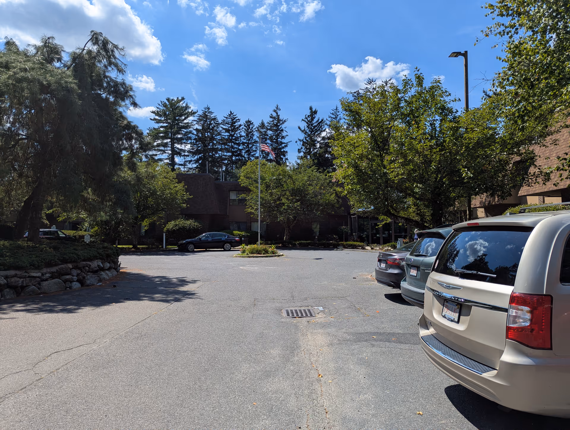 Sunny outdoor parking lot in front of a brick building, with trees and cars.