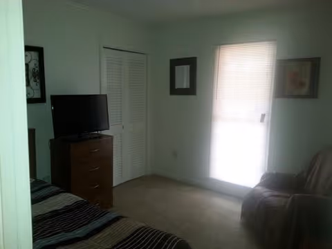 Small bedroom in a senior living facility with bed, dresser, TV, and window blinds.