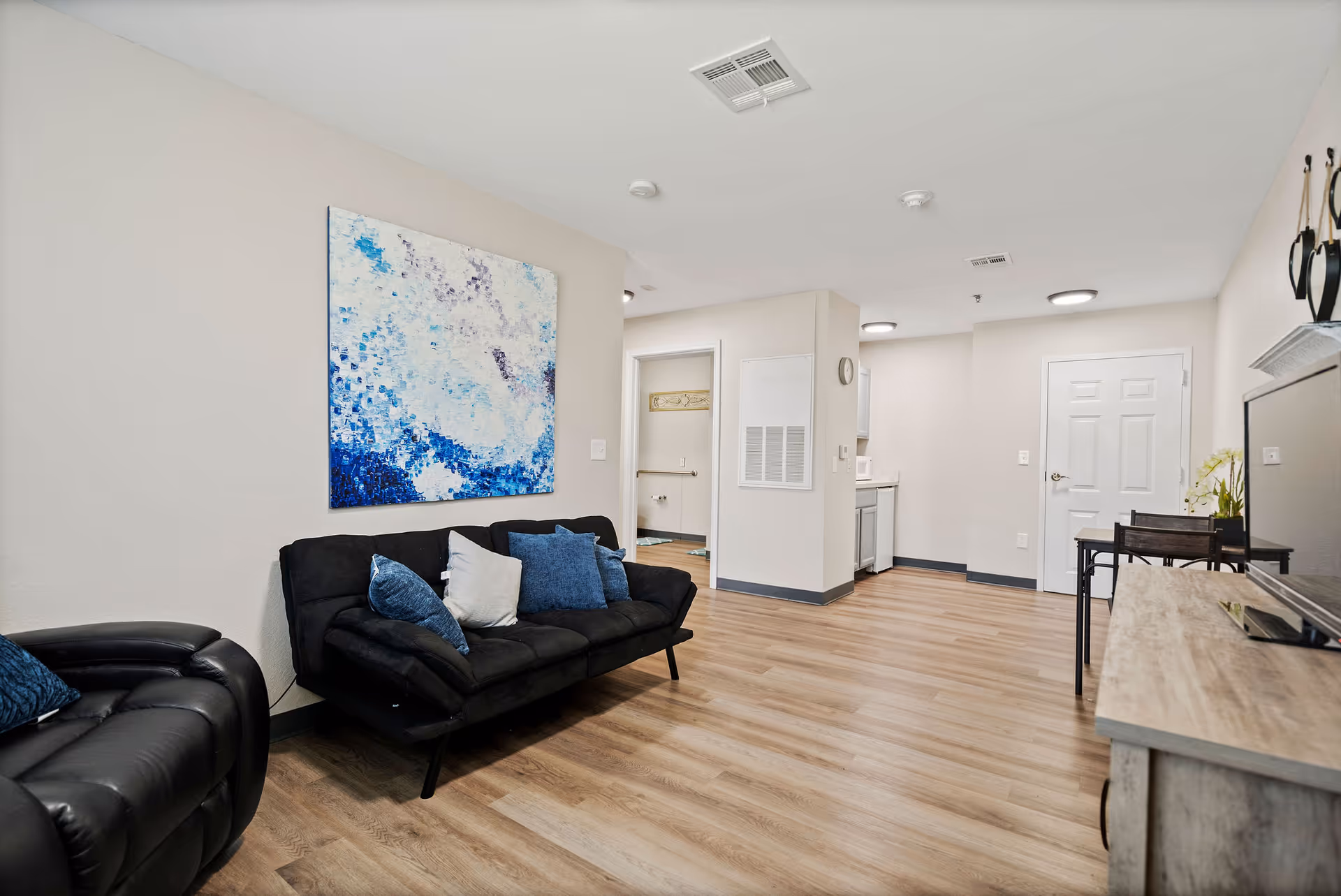Open living area with black sofas, abstract blue wall art, and a kitchenette.