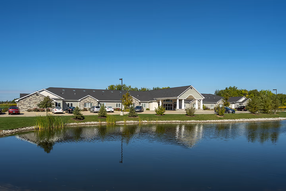 Exterior view of Villas of Holly Brook Assisted Living & Memory Care facility in Jerseyville, IL, showing a single-story building with a stone and siding facade, a covered entrance, several parked cars, and a pond with reflections in the foreground under a clear blue sky.