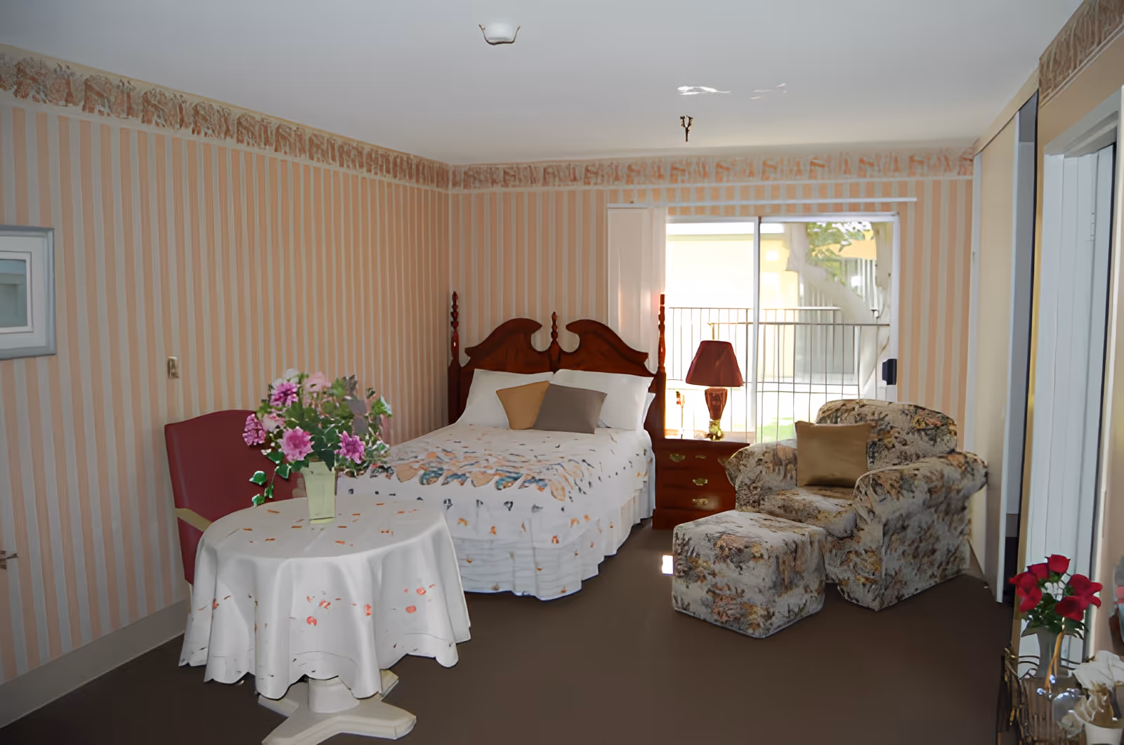 A cozy bedroom with a wooden bed featuring a white bedspread and two pillows. Next to the bed is a wooden nightstand with a red lamp. There is a floral upholstered armchair with a matching ottoman and a beige cushion. A small round table covered with a white tablecloth and a vase of pink flowers is positioned near a red chair. The room has striped wallpaper with a decorative border near the ceiling and a sliding glass door leading outside.