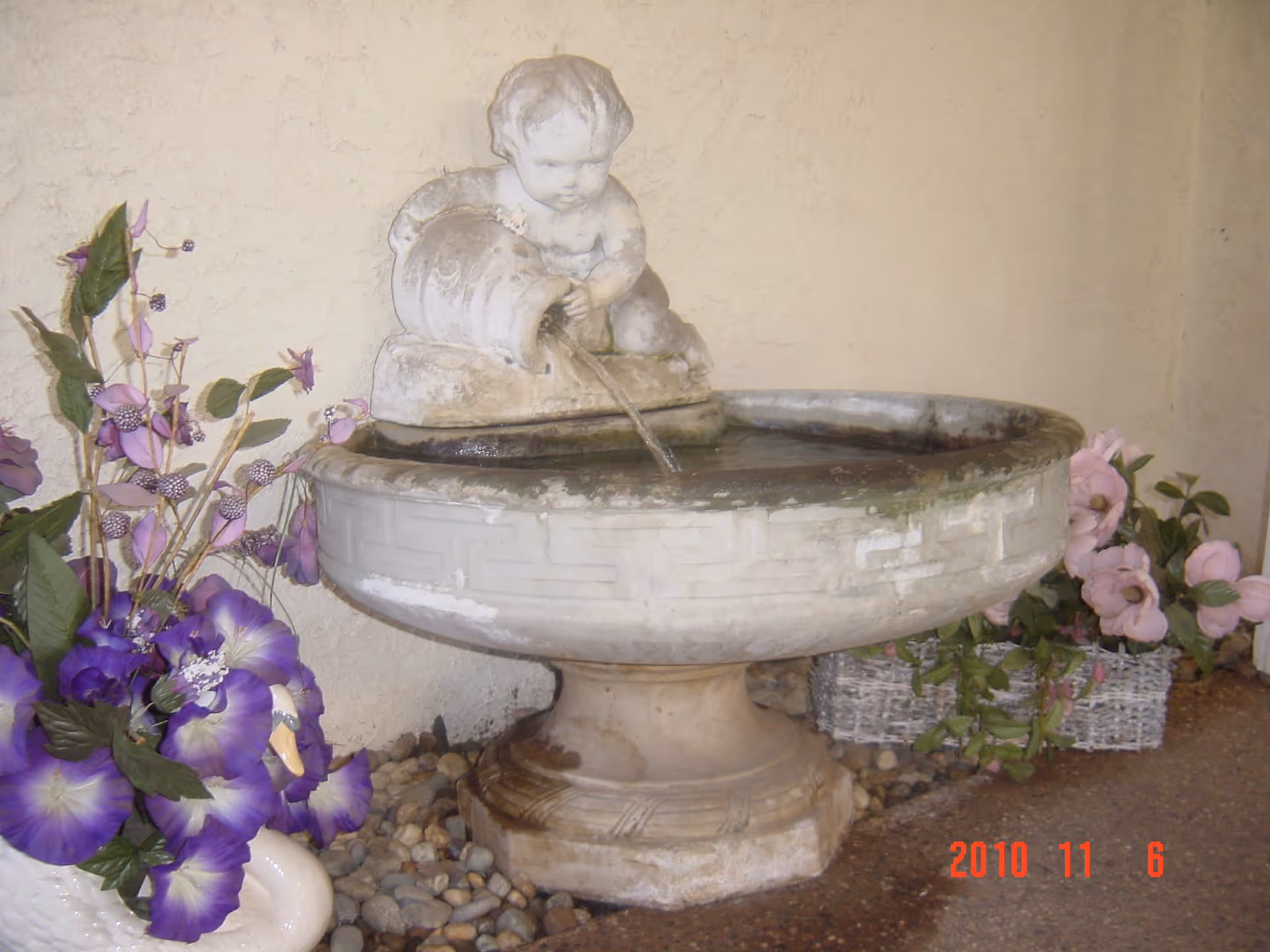 A decorative indoor water fountain featuring a statue of a child pouring water from a jug into a large basin, surrounded by artificial purple and pink flowers in baskets, set against a textured beige wall.