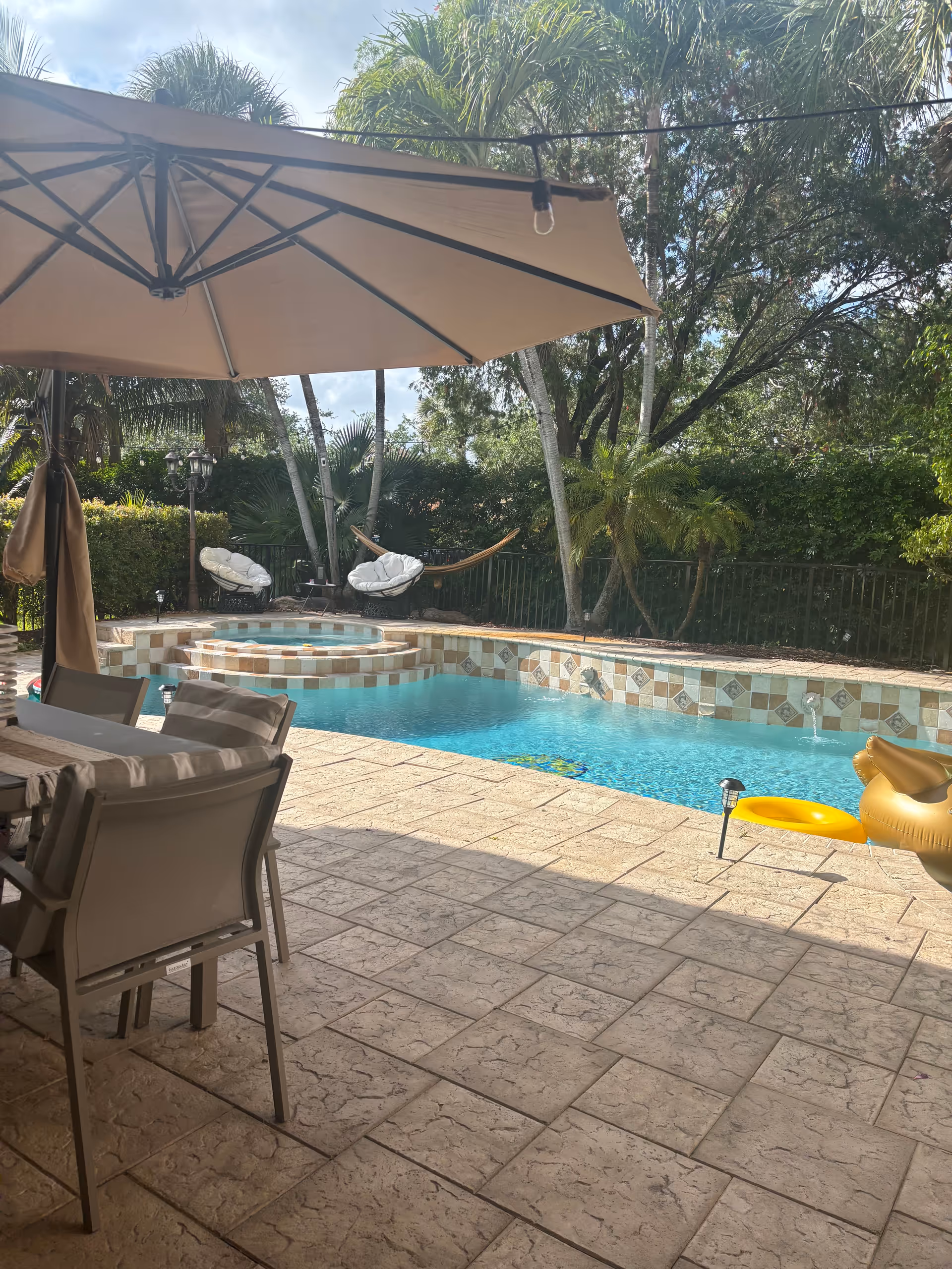 Outdoor pool area with seating, umbrellas, and palm trees surrounding a tiled pool.