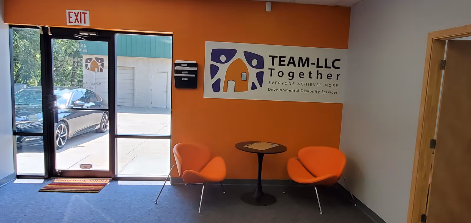 Orange-walled entrance lobby with two orange chairs, a small round table, near TEAM-LLC sign.
