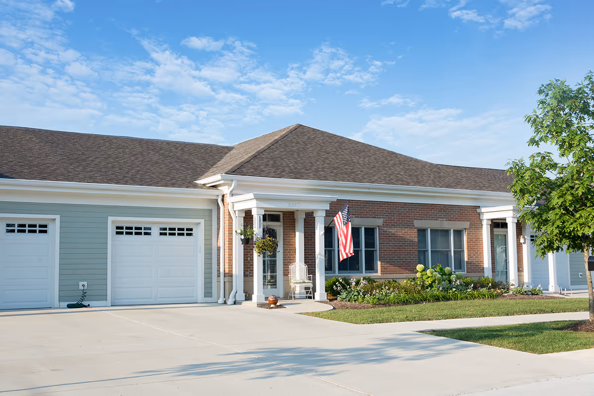 A single-story senior living building with brick and light siding, front porch, and American flag.