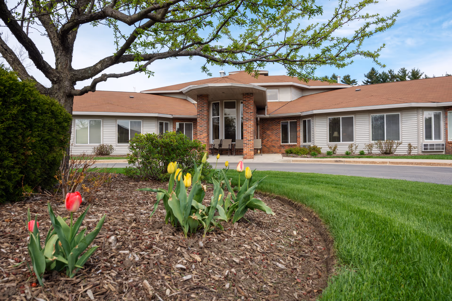 Front entrance of a single-story brick-and-siding senior living building with tulips and landscaped lawn in the foreground.